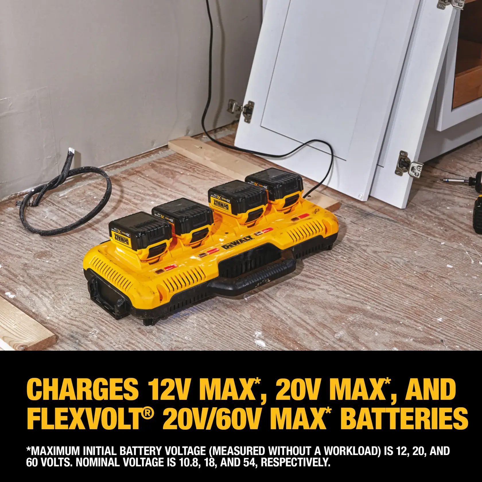 A yellow DEWALT four-port battery charger on a wooden floor, charging four DEWALT batteries. The charger supports 12V Max, 20V Max, and FLEXVOLT 20V/60V Max batteries. Nearby are construction materials and a white cabinet door.