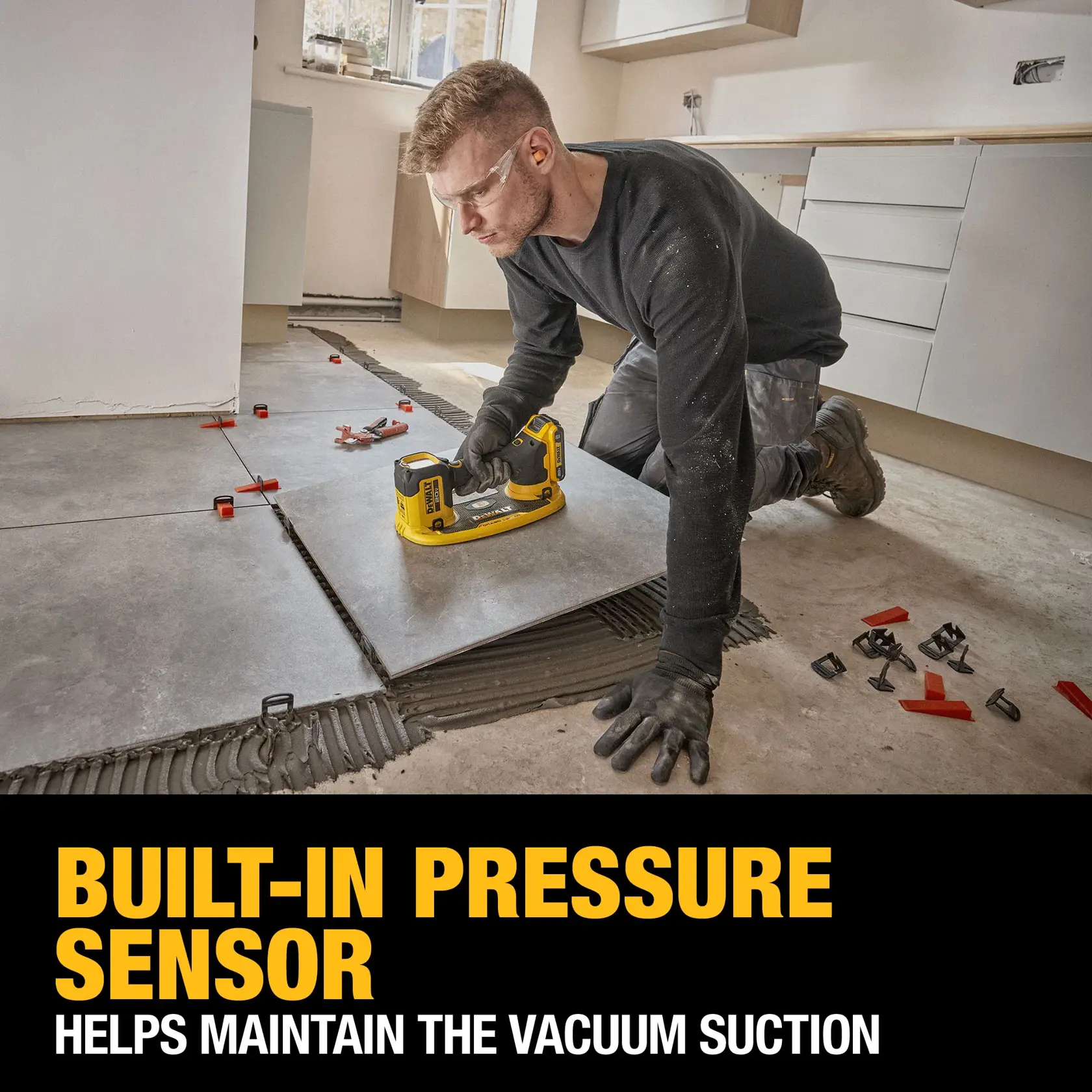 A person is installing floor tiles using a DEWALT tool, featuring a built-in pressure sensor for maintaining vacuum suction. The scene includes large gray tiles, installation clips, and the person is kneeling on the floor wearing gloves. Text on the image highlights the product feature.