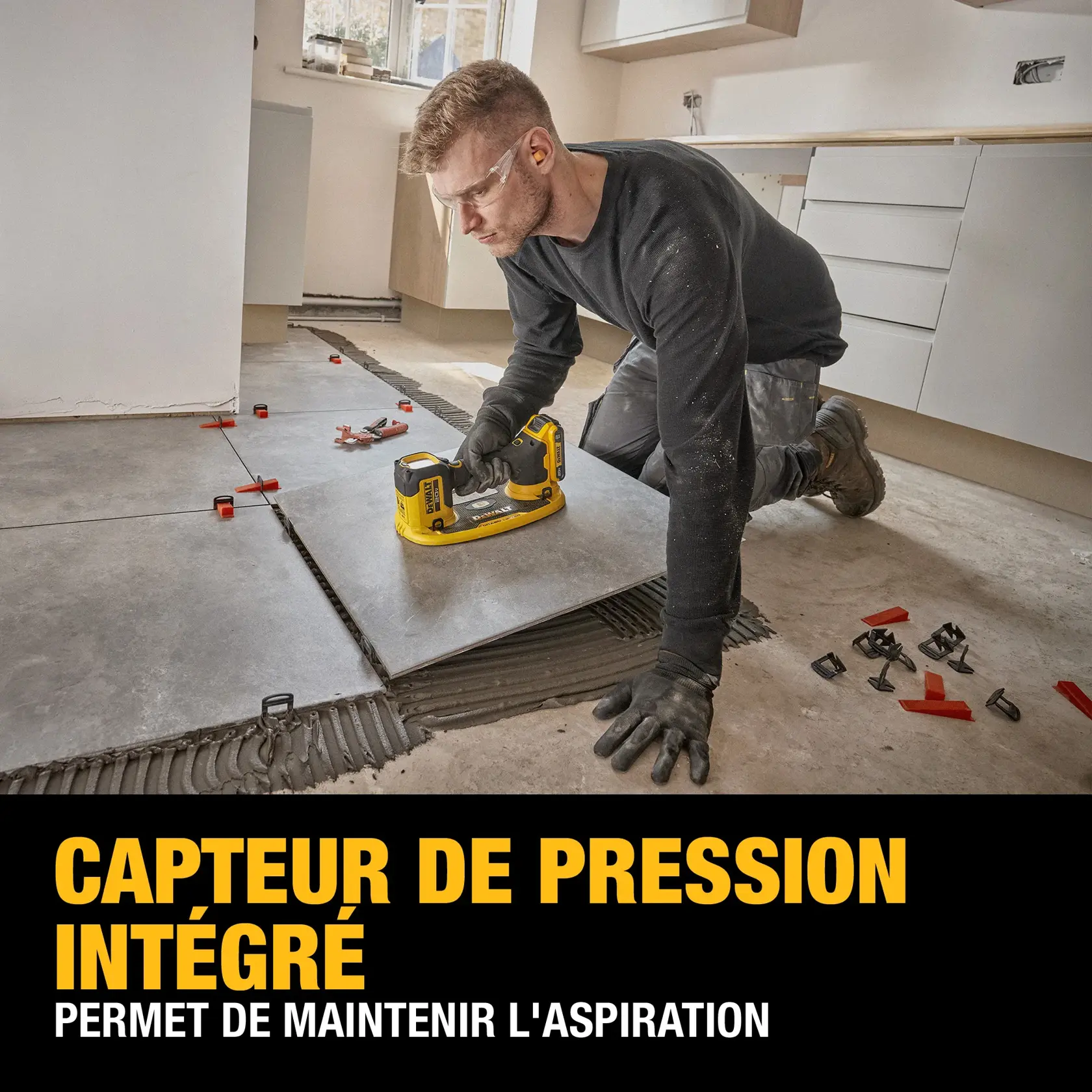 A person is installing floor tiles using a DEWALT tool, featuring a built-in pressure sensor for maintaining vacuum suction. The scene includes large gray tiles, installation clips, and the person is kneeling on the floor wearing gloves. Text on the image highlights the product feature.