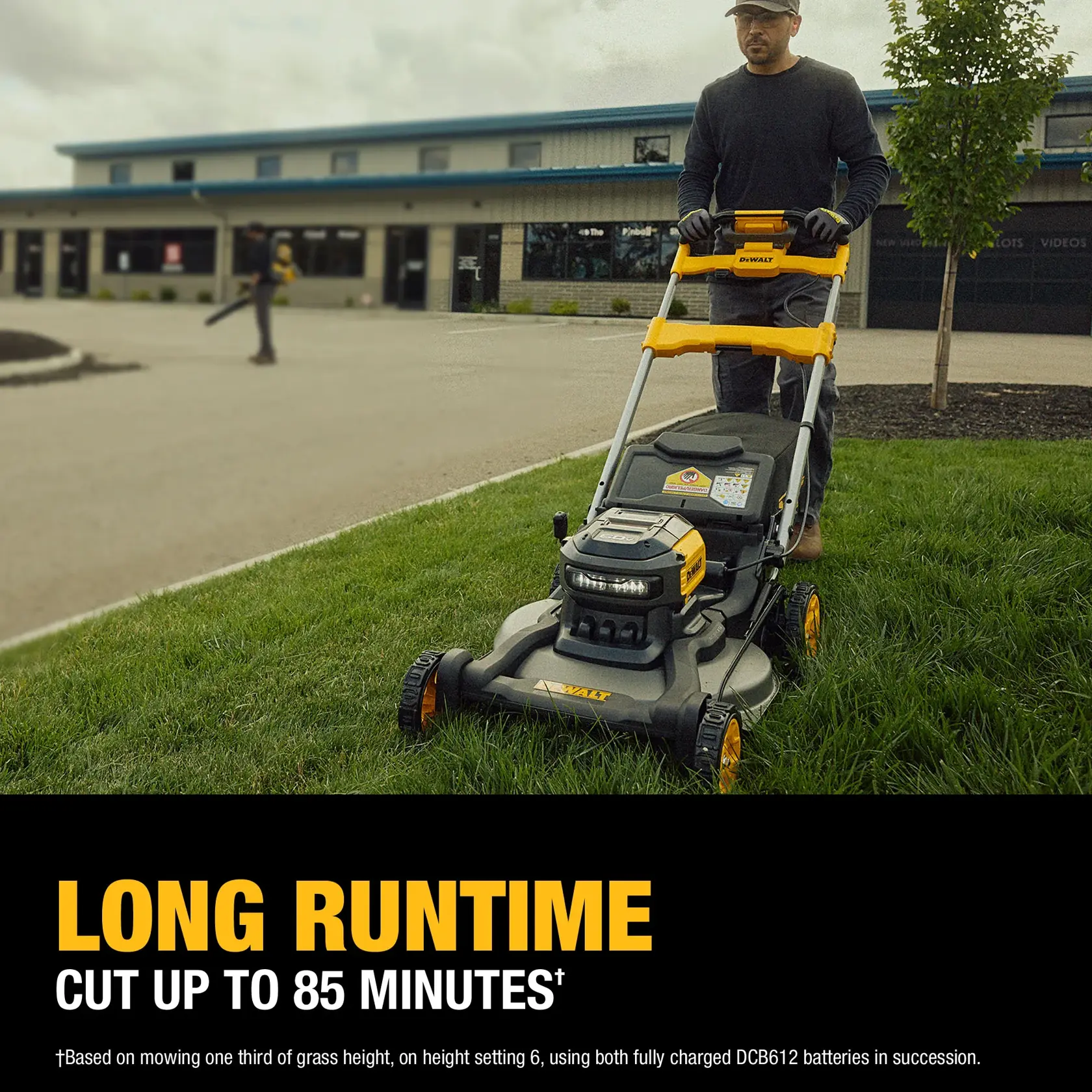 A person is pushing a DEWALT battery-powered lawn mower on a grassy area near a building. The mower features a yellow and black design and the text on the image highlights its long runtime of up to 85 minutes.