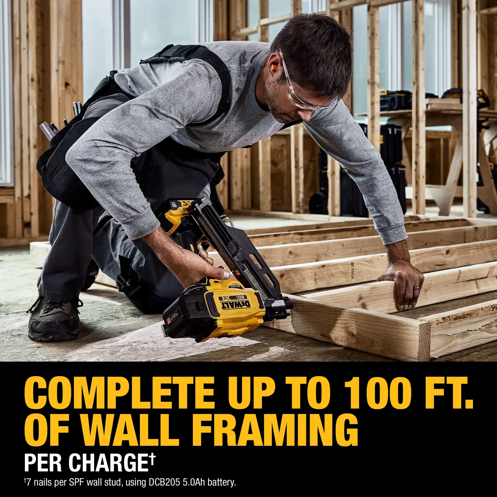 A person in protective glasses kneels in a construction area, using a DEWALT DCN930P1 cordless nailer to fasten wooden boards for wall framing. The image highlights the tool's capability to complete up to 100 feet of wall framing per battery charge. Construction materials and unfinished wooden framing are visible in the background.
