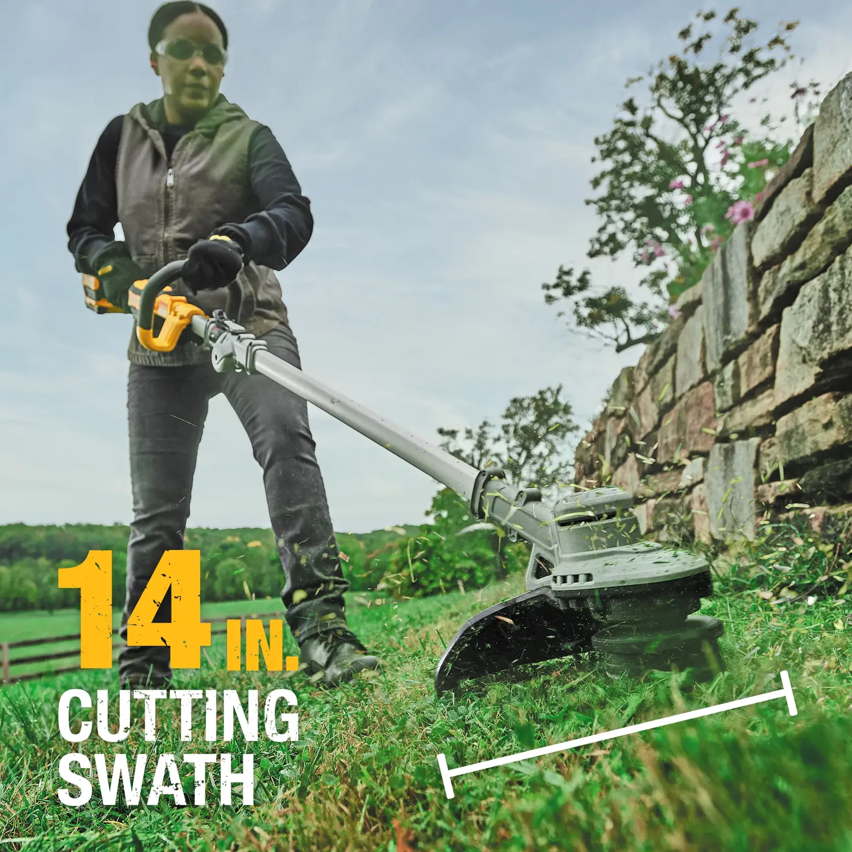 Close-up of a person using a DEWALT string trimmer to cut grass near a stone wall.
