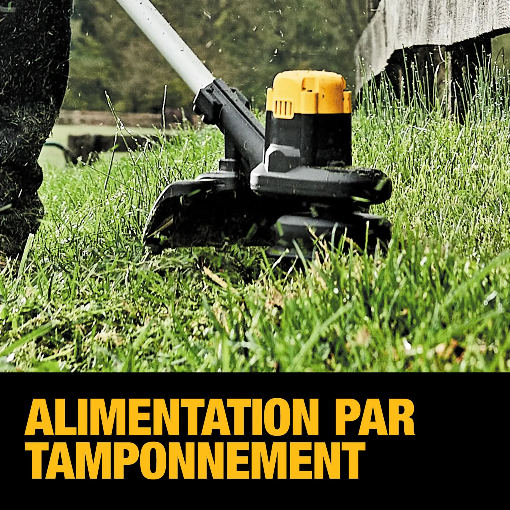 A person using a DEWALT cordless string trimmer (model DCST925B) to trim grass along a stone wall in a garden or outdoor area. The individual is wearing protective gloves and dark clothing. The landscape features green grass, trees, and flowers growing above the wall.