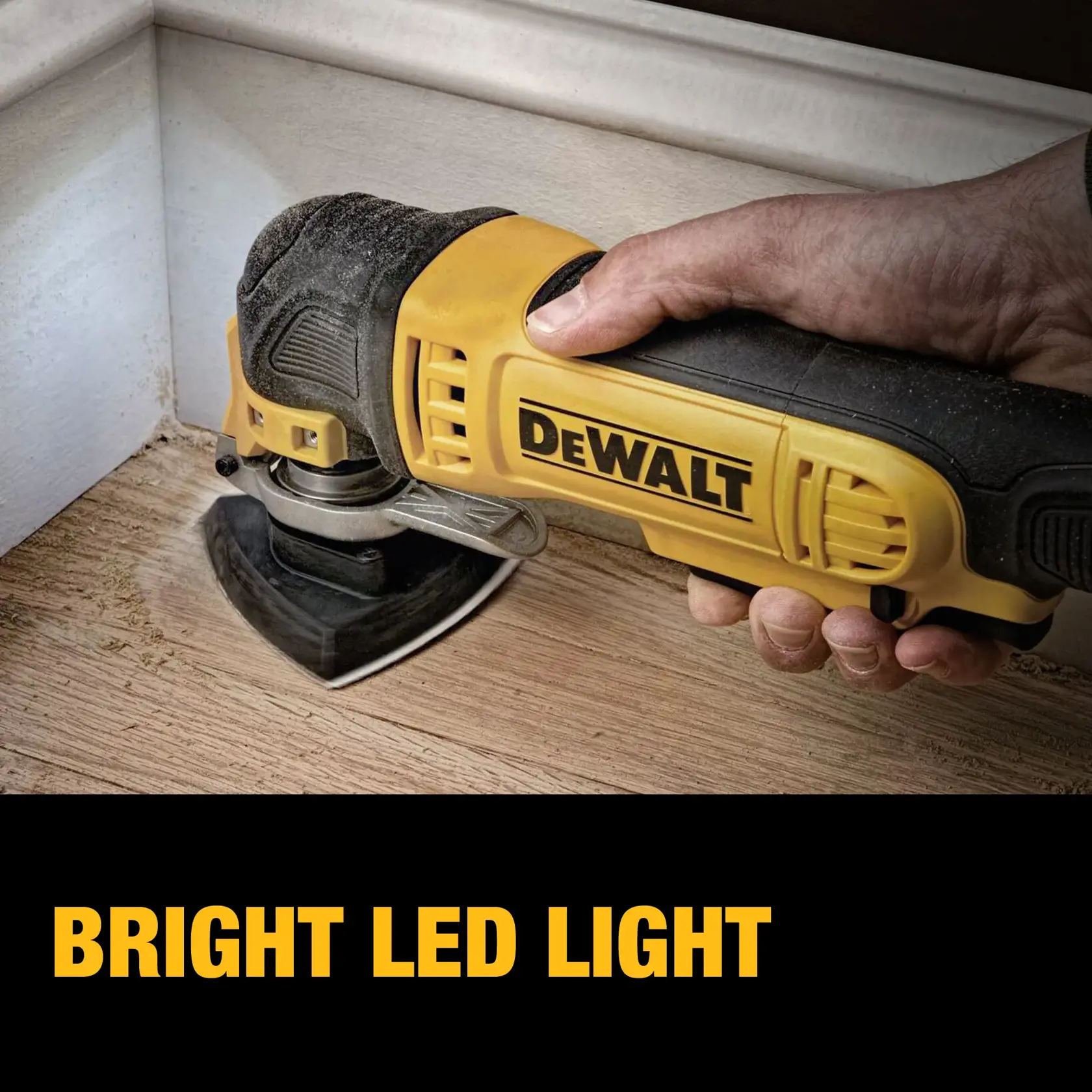 A hand holds a DEWALT DWE315 oscillating multi-tool, sanding wood flooring near the edge of a baseboard. The bright LED light is emphasized in the image.