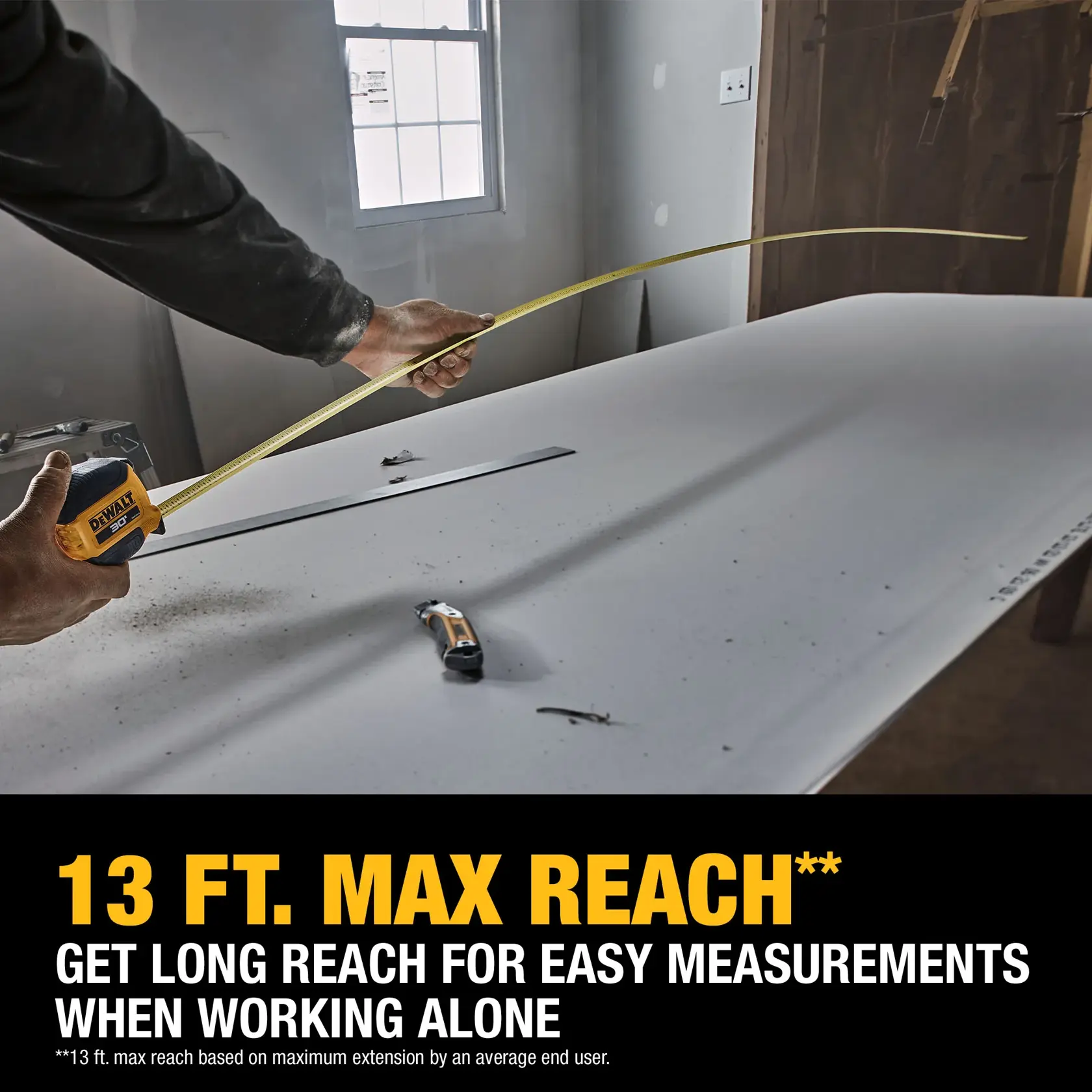 A person uses a DEWALT tape measure to measure a long piece of material on a work table in a construction setting. The tape is extended to show the 13 ft. maximum reach feature for easy measurements when working alone. Promotional text highlighting the long reach capability is visible at the bottom.