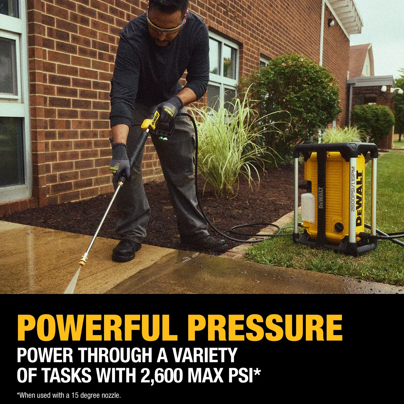 A person using a DEWALT DWPW2600 pressure washer to clean a concrete walkway outside a brick building. The washer is shown spraying water with strong pressure. The text highlights 'Powerful Pressure' with 2,600 max PSI output, and there is a yellow DEWALT pressure washer unit visible on the grass.