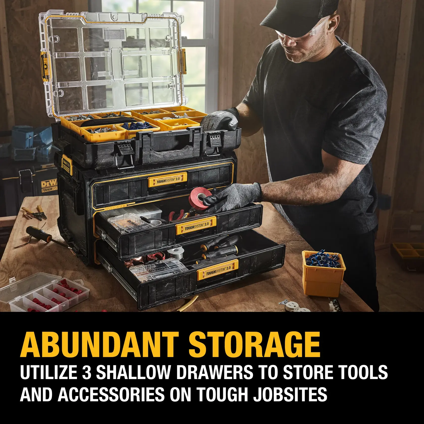A person uses a DEWALT ToughSystem 2.0 storage unit with three shallow drawers and an open top compartment, organizing tools and accessories on a workbench in a workshop.