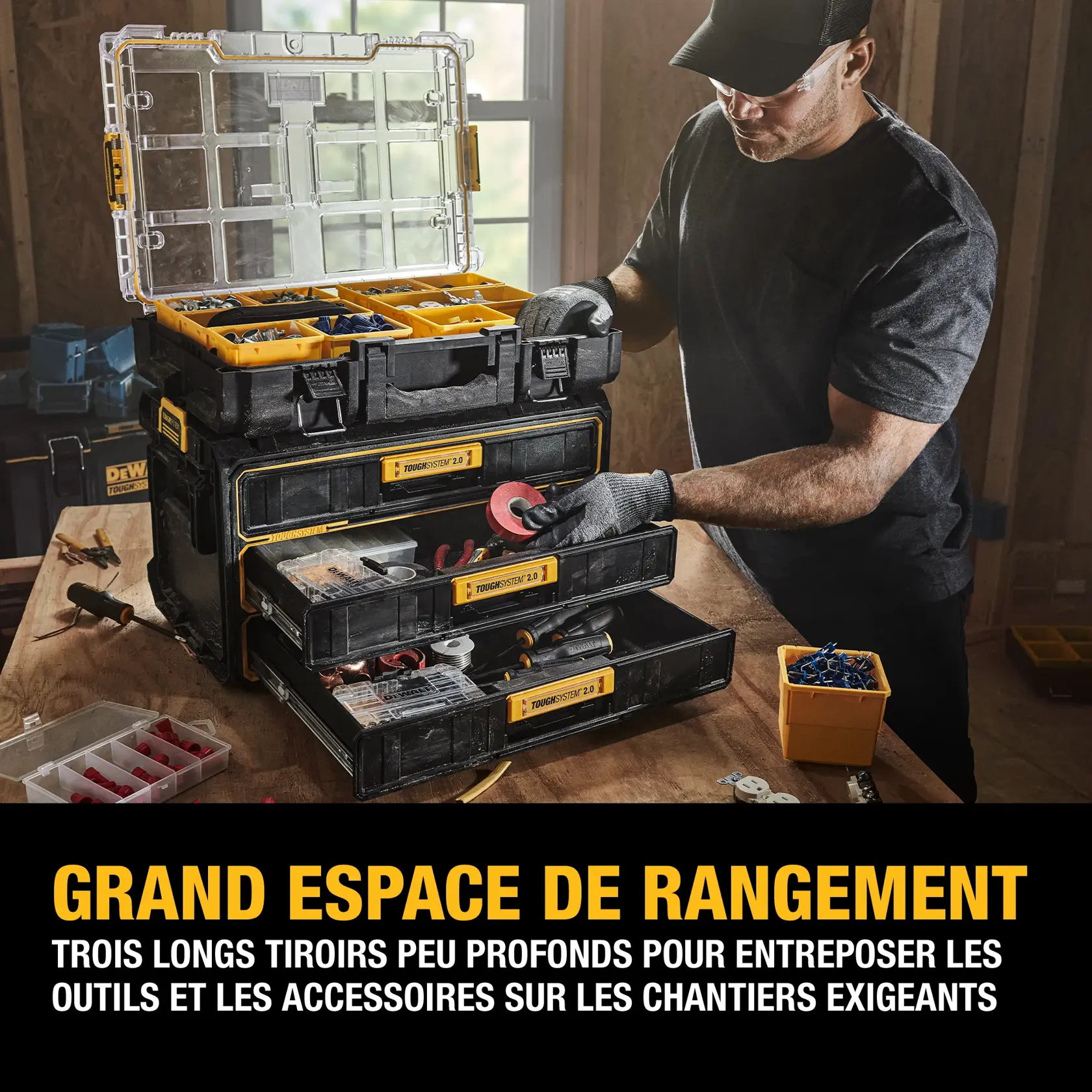 A person wearing work gloves is organizing tools and accessories in a DEWALT ToughSystem 2.0 storage system with three shallow drawers and a clear-lid top compartment, placed on a workbench in a workshop setting. The bottom of the image features French text promoting large storage space and the shallow drawers for tool organization on demanding job sites.
