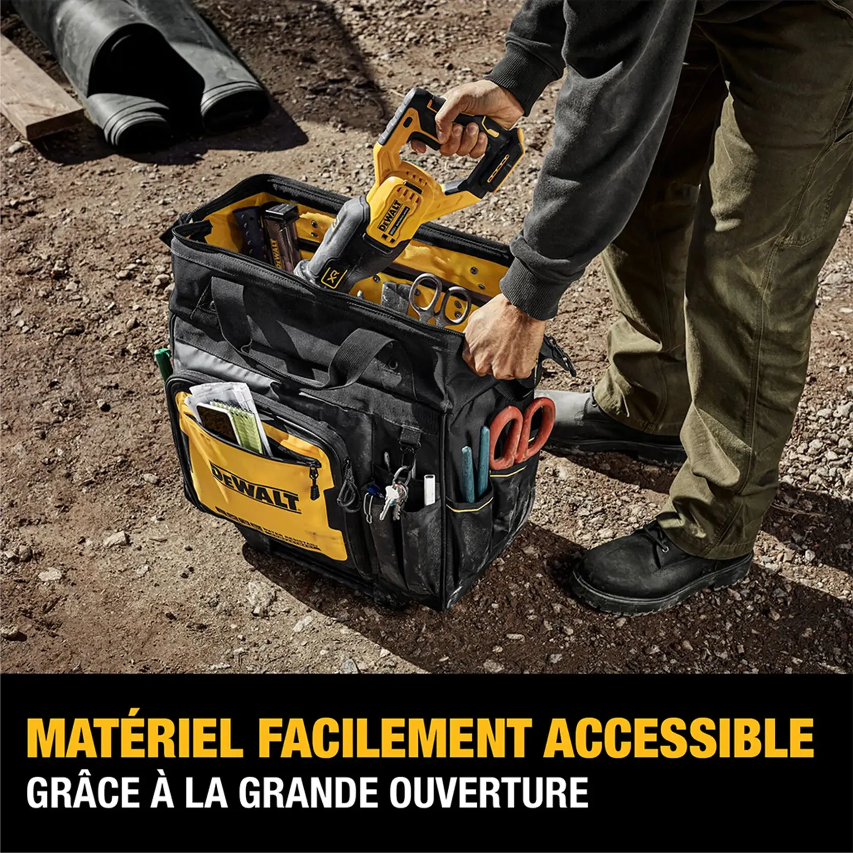 A person stands on a gravel surface and opens a DEWALT tool bag, showing its large opening and organized compartments filled with various tools, including a cordless reciprocating saw, pliers, and markers. The bag highlights easy access to tools.