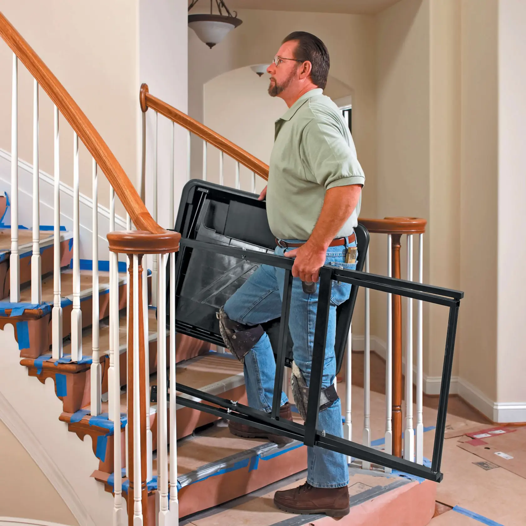 A person with a blurred face is carrying a large black frame up a staircase in a residential setting. The stairs and surrounding area are covered with protective materials, indicating construction or renovation work.