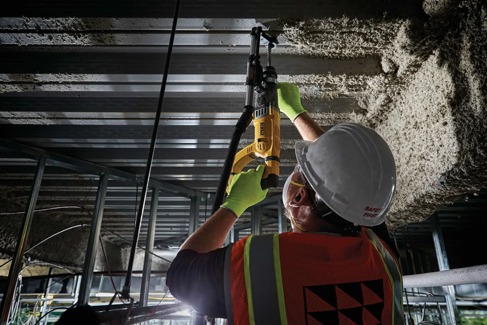 A worker wearing a hard hat, safety vest, gloves, and protective eyewear is using a yellow DEWALT rotary hammer drill to work on a ceiling in a construction setting.