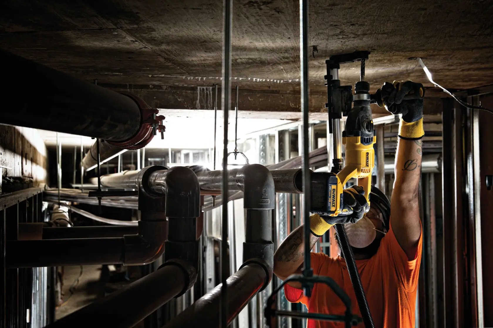 A worker wearing gloves and an orange shirt uses a yellow DEWALT power tool to drill into a concrete ceiling in an industrial setting, surrounded by multiple pipes and metal structures.