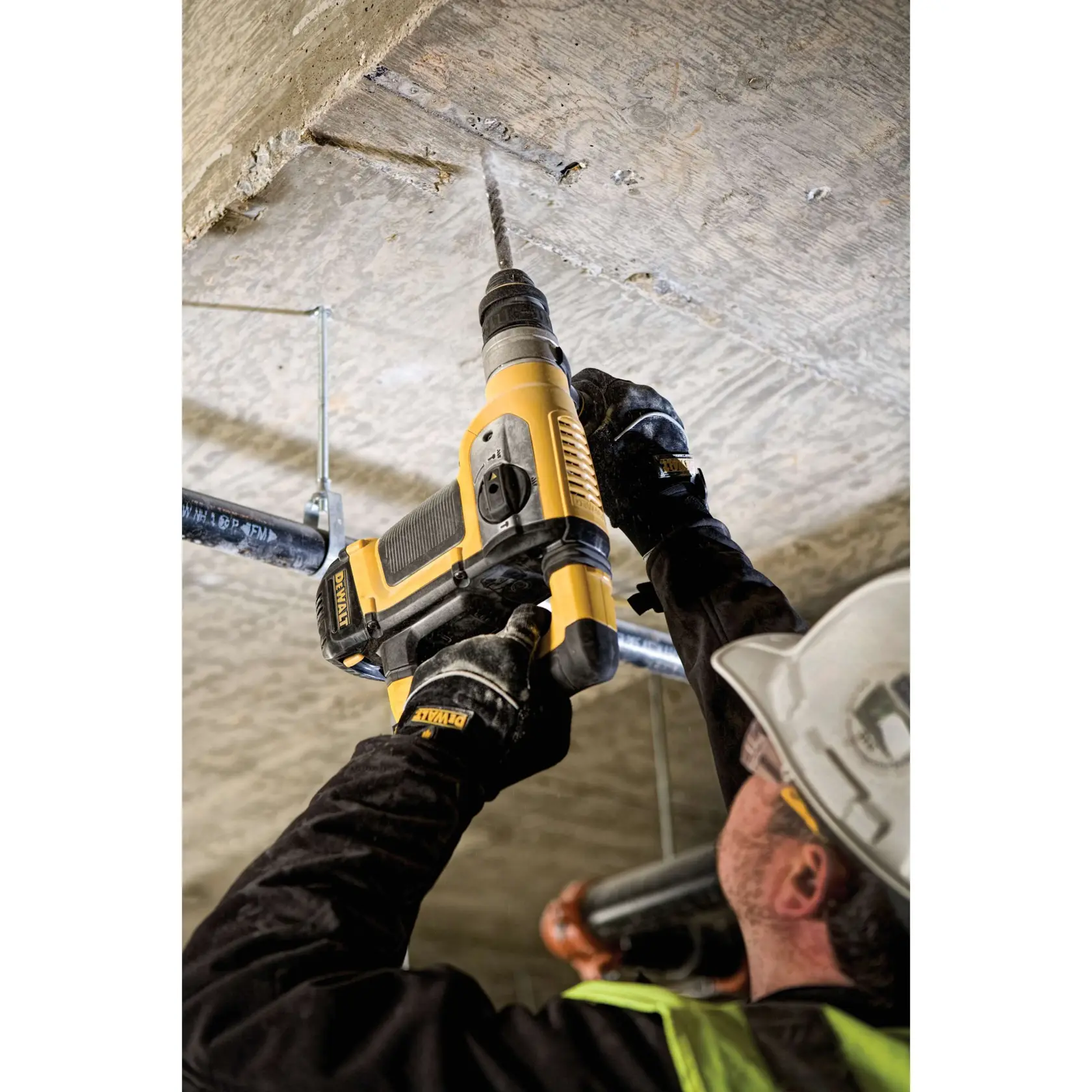 A person wearing a hard hat and safety gloves using a yellow DEWALT rotary hammer drill to drill into a concrete ceiling on a construction site.