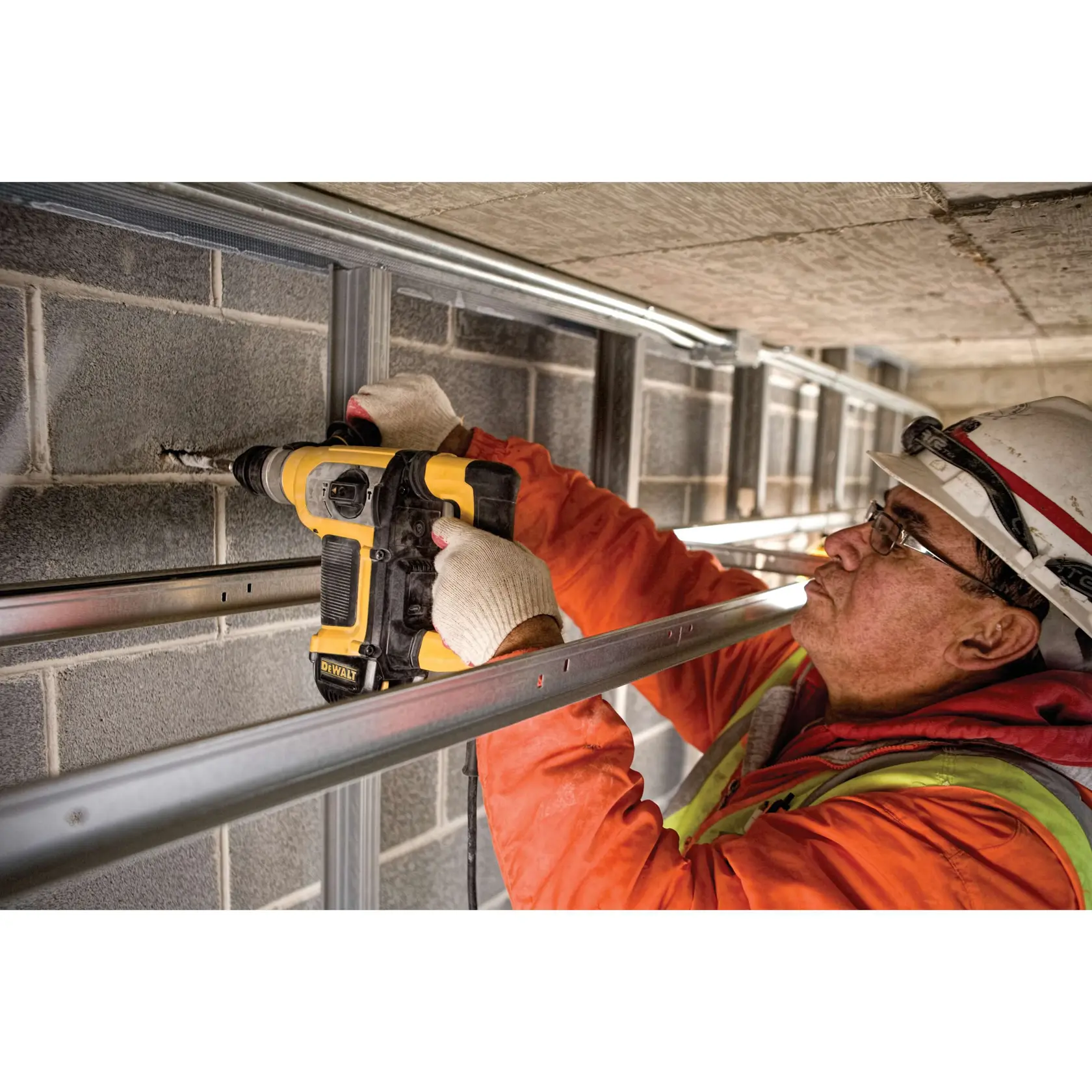 A construction worker wearing gloves and safety gear uses a yellow and black DEWALT rotary hammer drill to drill into a concrete block wall.