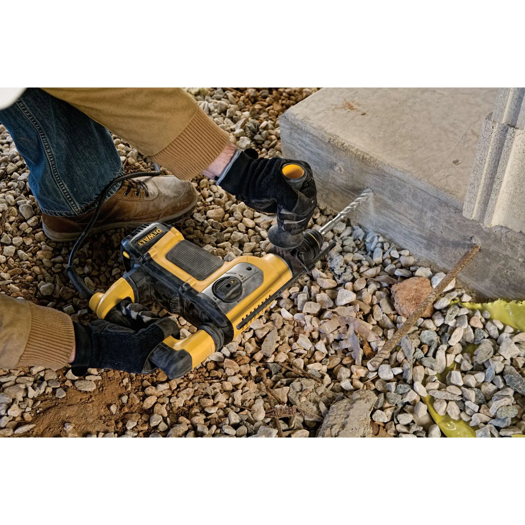 A person wearing gloves and work boots uses a yellow and black DEWALT hammer drill to drill into a concrete block surrounded by gravel.