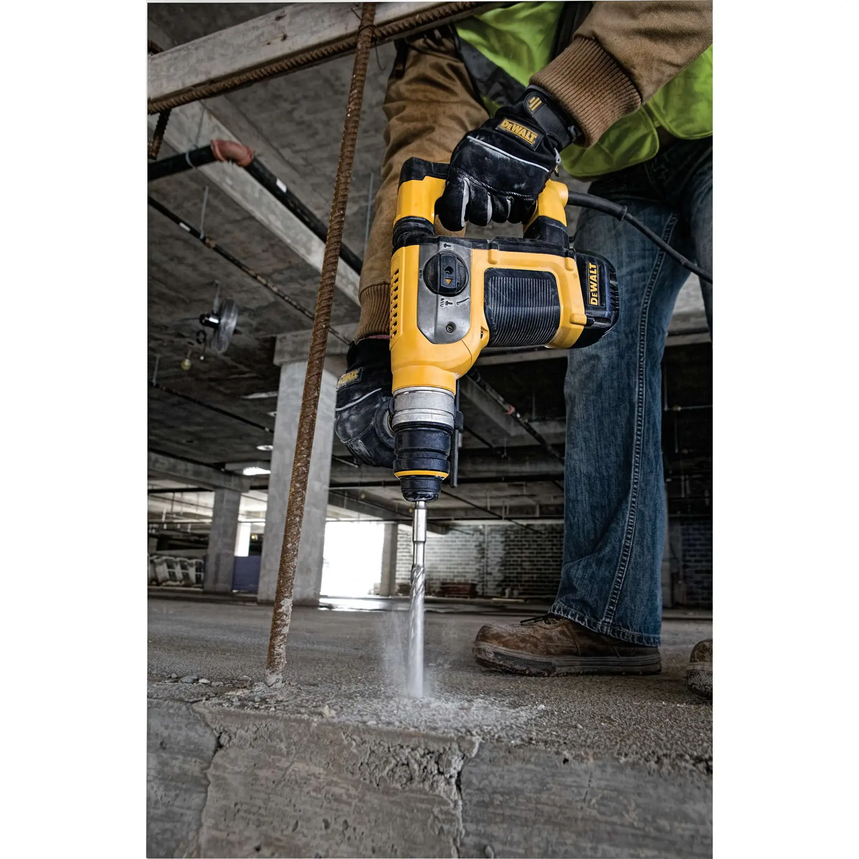 A worker uses a DEWALT power tool to drill into a concrete floor at a construction site. The tool is yellow and black and is producing dust as it works.