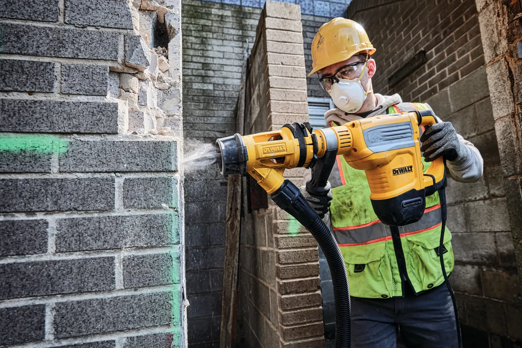 A construction worker wearing a yellow hard hat and safety vest uses a DEWALT rotary hammer drill (model D25614K) to break through a brick wall, with dust being expelled through an attached dust extraction system.