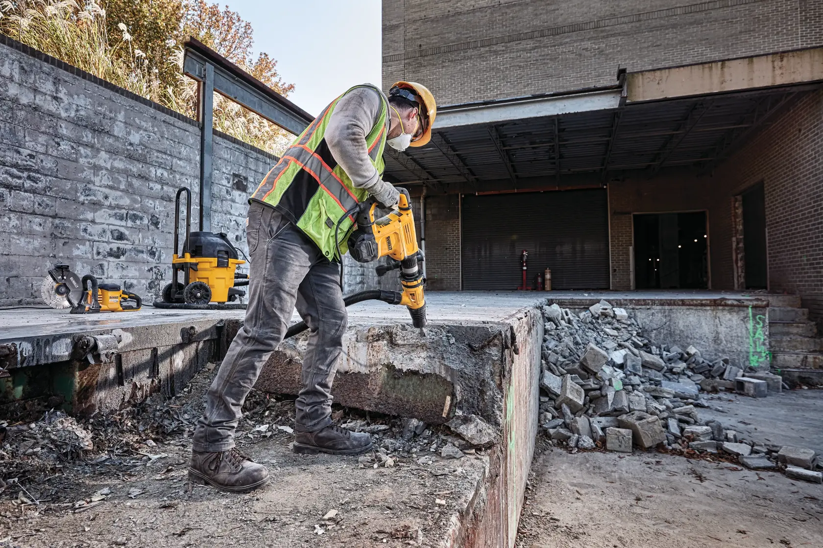 A construction worker wearing safety gear uses a DEWALT demolition hammer on a concrete structure at a worksite. Crumbled concrete and debris are visible around the area.