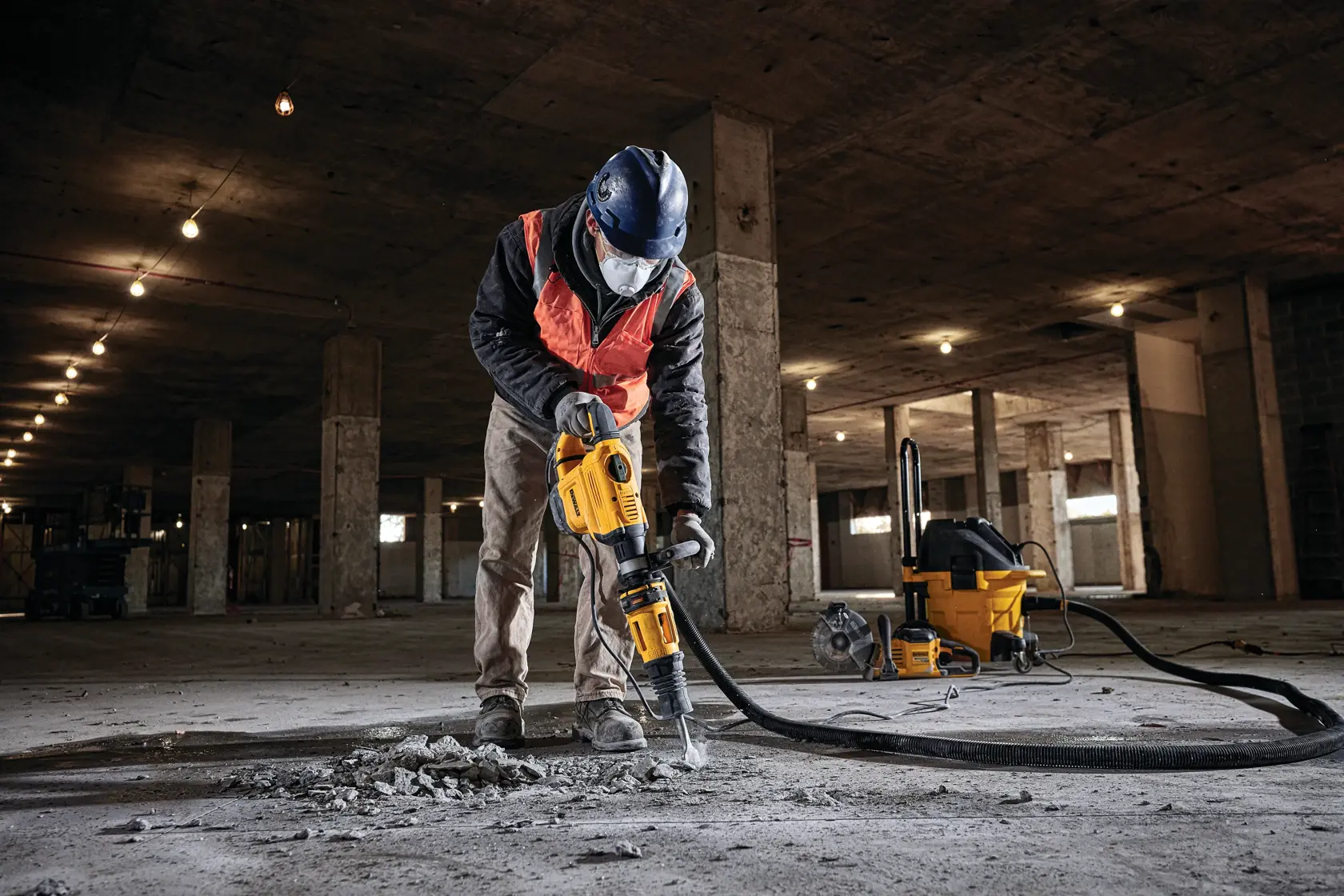 A person wearing safety gear, including a hard hat and face mask, uses a DEWALT yellow demolition hammer to break concrete on the floor of a large construction site. A yellow DEWALT vacuum machine is visible in the background.