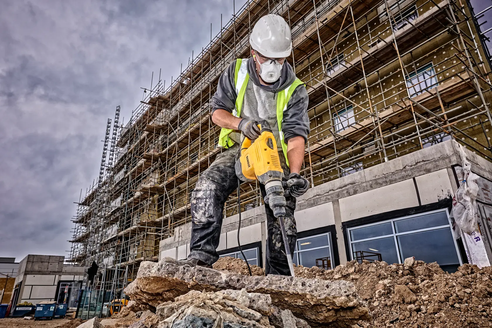 A construction worker wearing a safety helmet and high-visibility vest is using a yellow DEWALT demolition hammer to break concrete at a construction site. Scaffolding and an unfinished building are visible in the background.