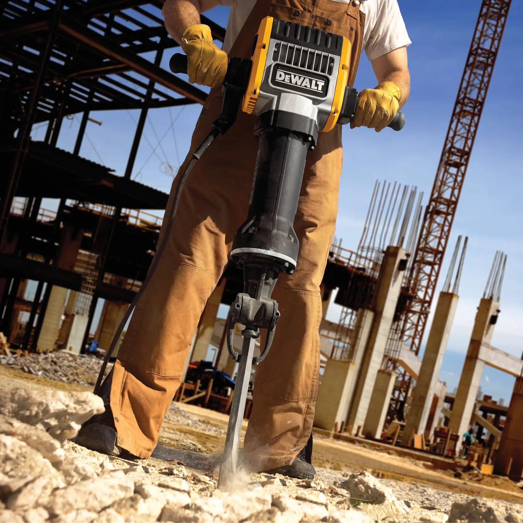 A person wearing work gloves and overalls operates a DEWALT jackhammer, breaking concrete at a construction site with structural steel and columns visible in the background.