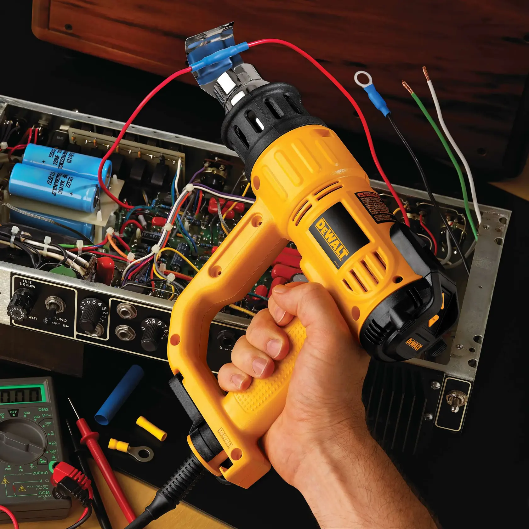 A person is using a DEWALT D26960K heat gun on an electronic component, likely heat shrinking tubing over electrical wires while working on an open circuit board. Various tools, wires, and a multimeter are visible around the work area.
