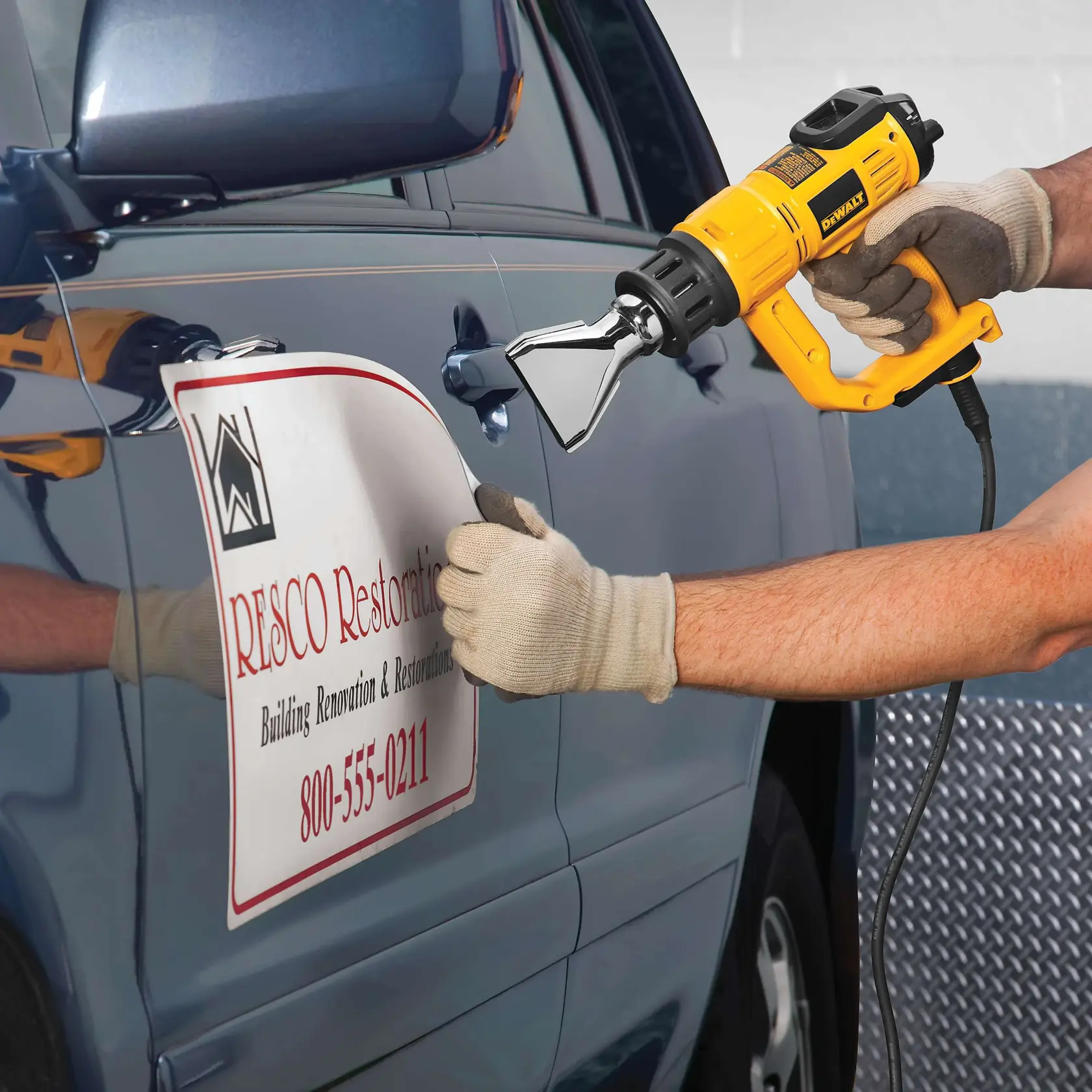 A person wearing gloves uses a yellow DEWALT heat gun (model D26960K) to remove a business decal from the side of a blue car.