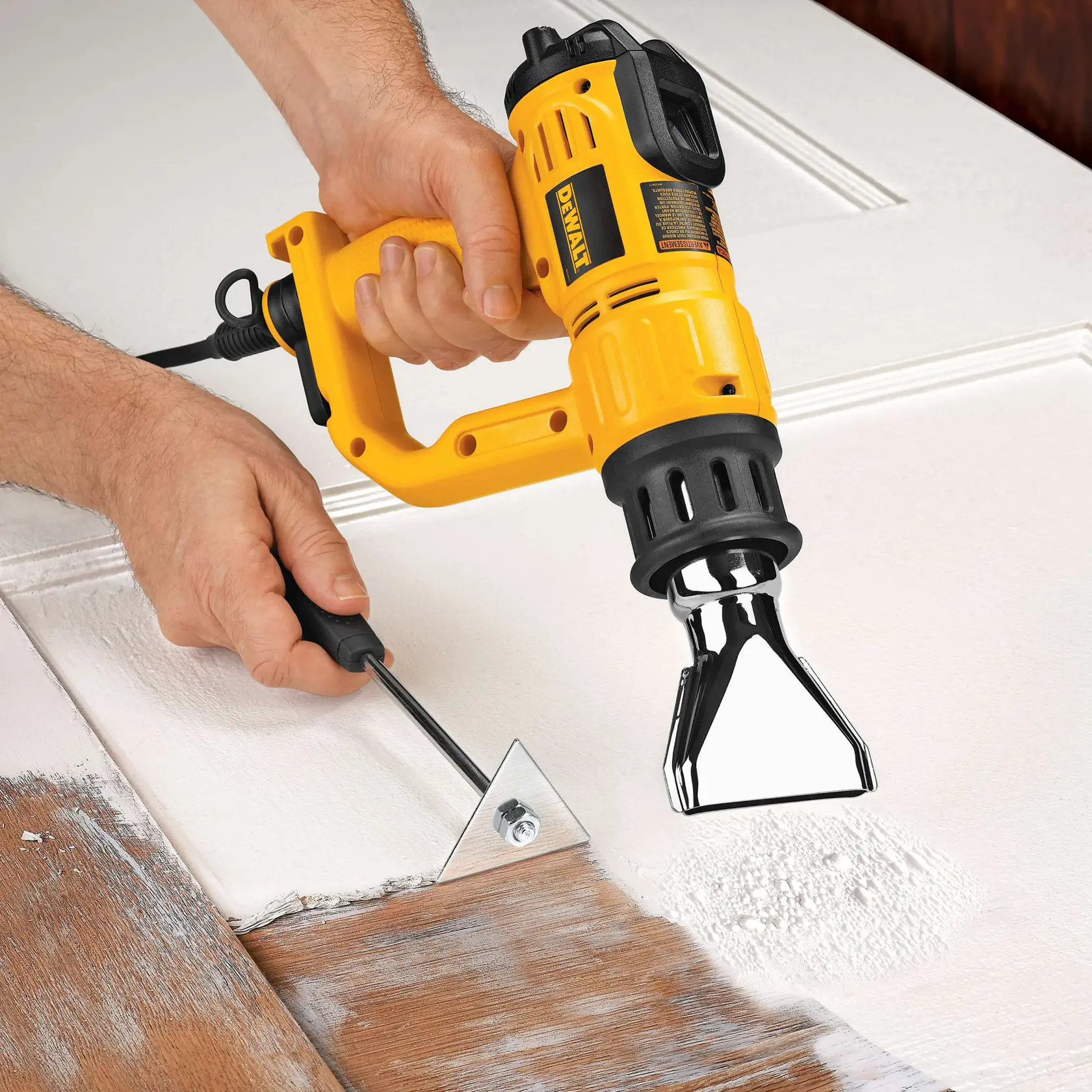 A person uses a yellow DEWALT heat gun and a metal scraper to remove paint from a wooden surface.