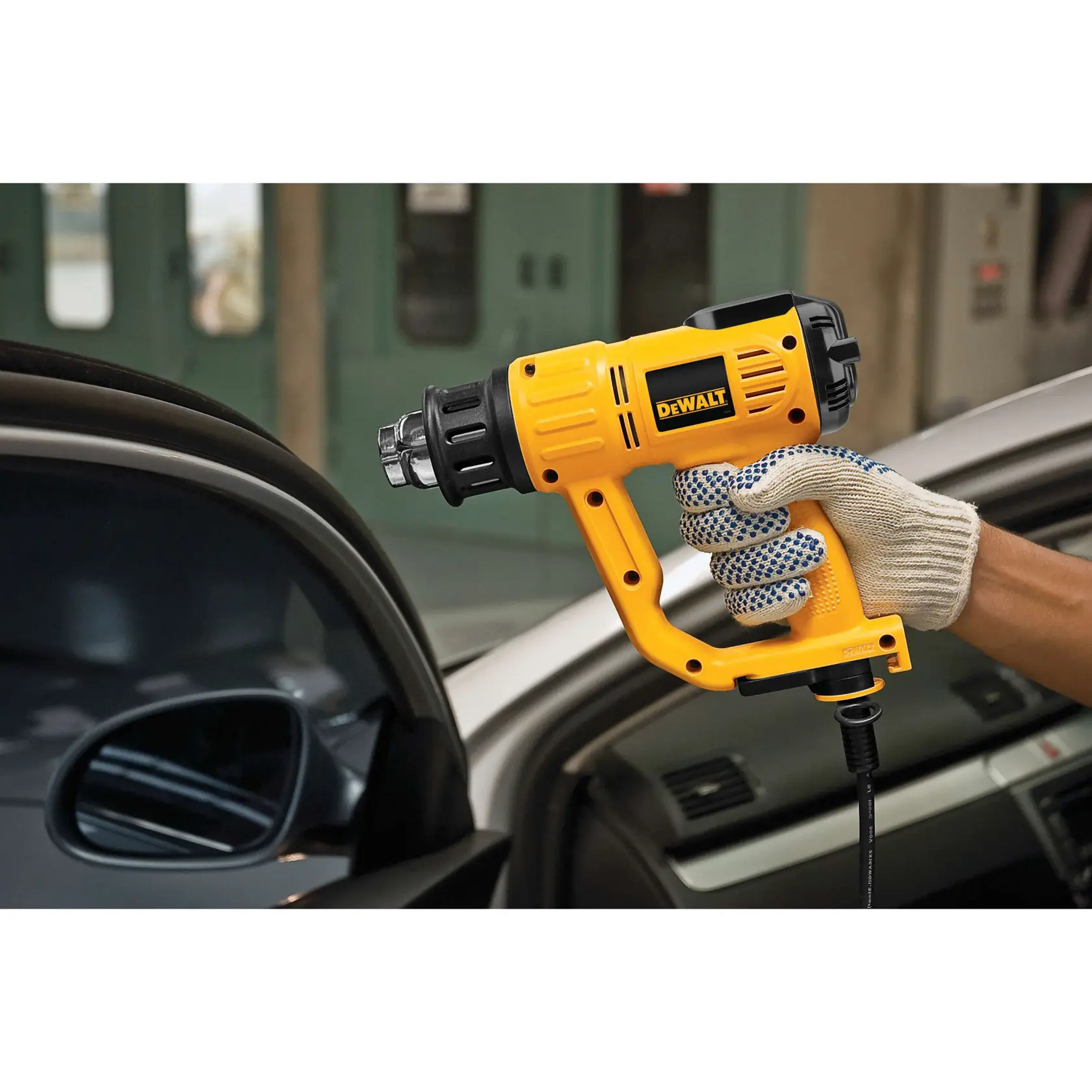 A person wearing a work glove is using a yellow DEWALT heat gun near the window of a car.