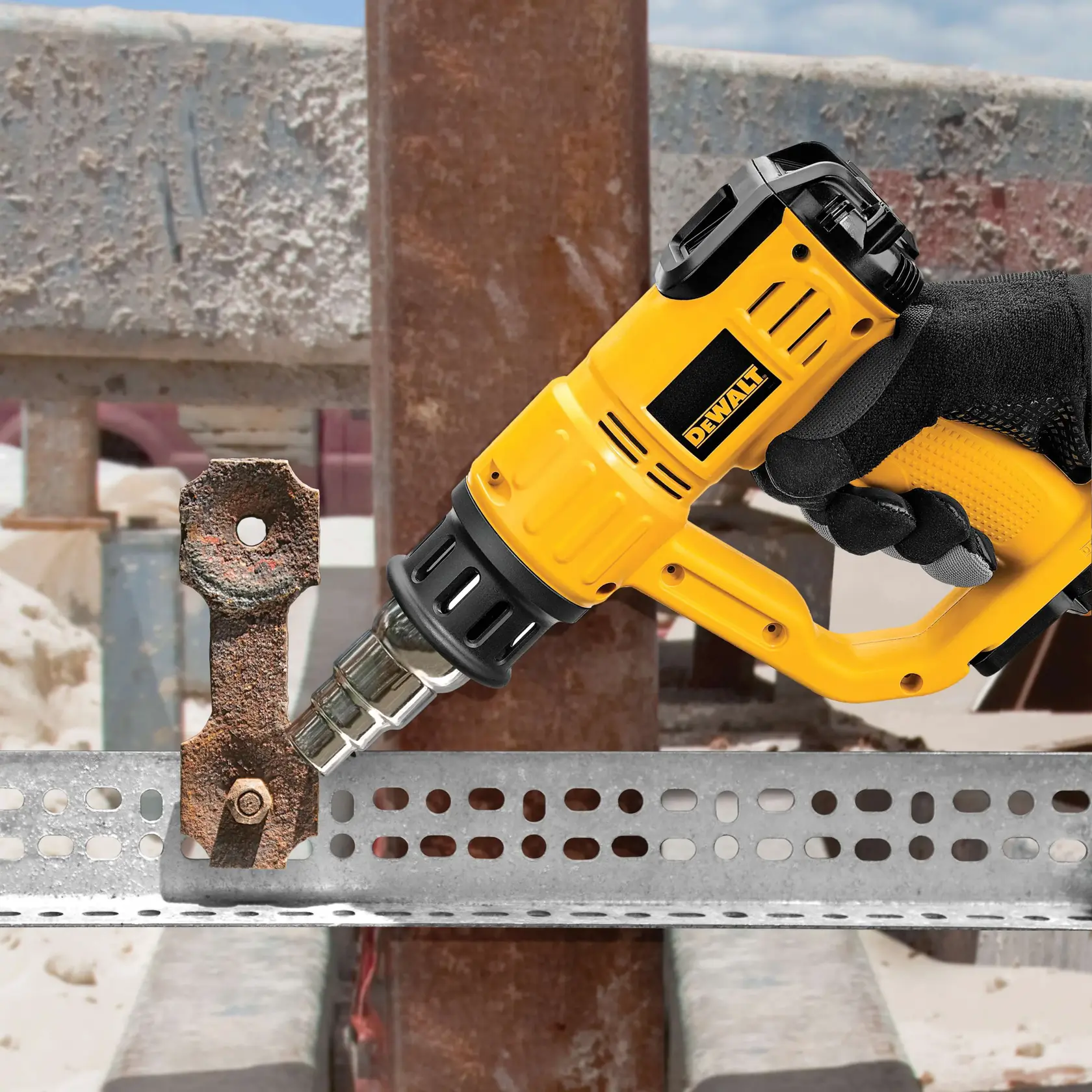 A hand wearing a black glove holds a yellow DEWALT heat gun (model D26960) pointed at a rusty metal bracket attached to a perforated metal beam, with a construction site in the background.