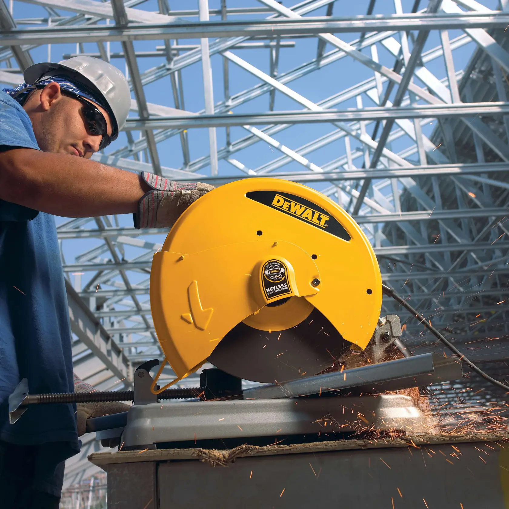 A person wearing work gloves and a hard hat operates a DEWALT yellow chop saw to cut metal, with sparks flying, inside a building with steel framework.