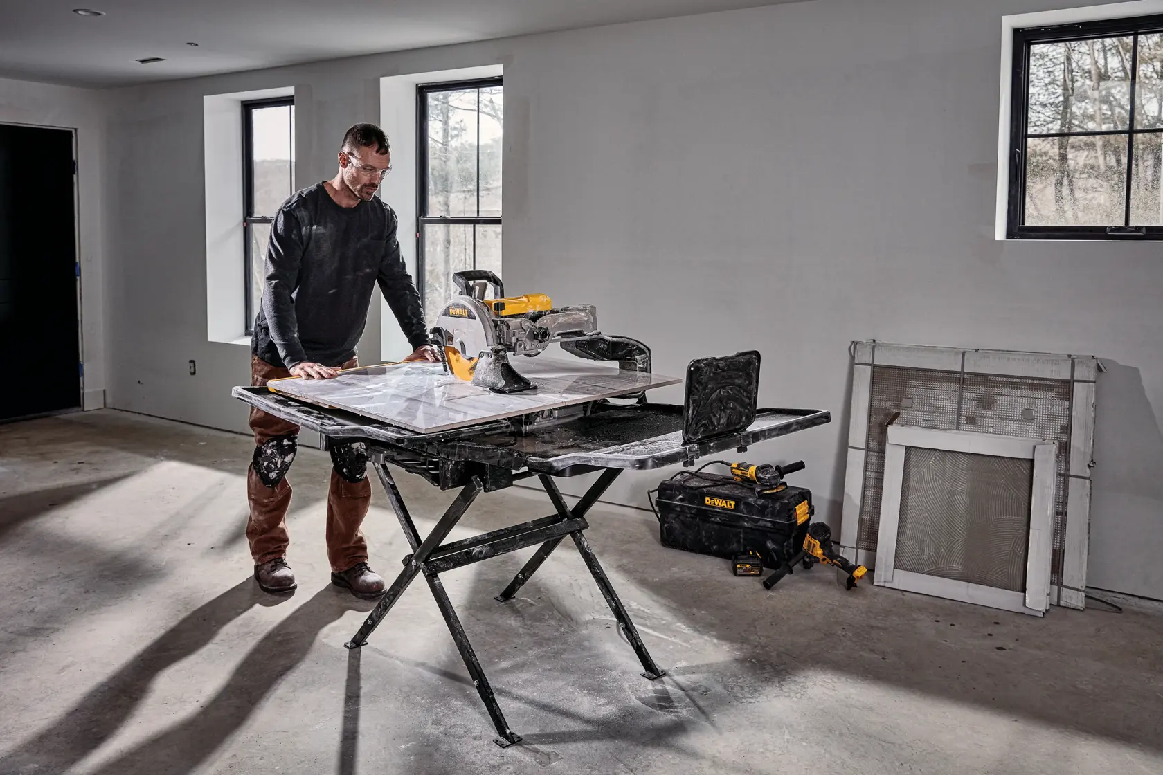 A person stands beside a DEWALT D36000S tile saw setup on its stand in a room under construction. The saw is placed on a foldable black stand, and there are tools and a DEWALT tool bag on the floor nearby. The room has unfinished walls, windows, and construction materials leaning against the wall.