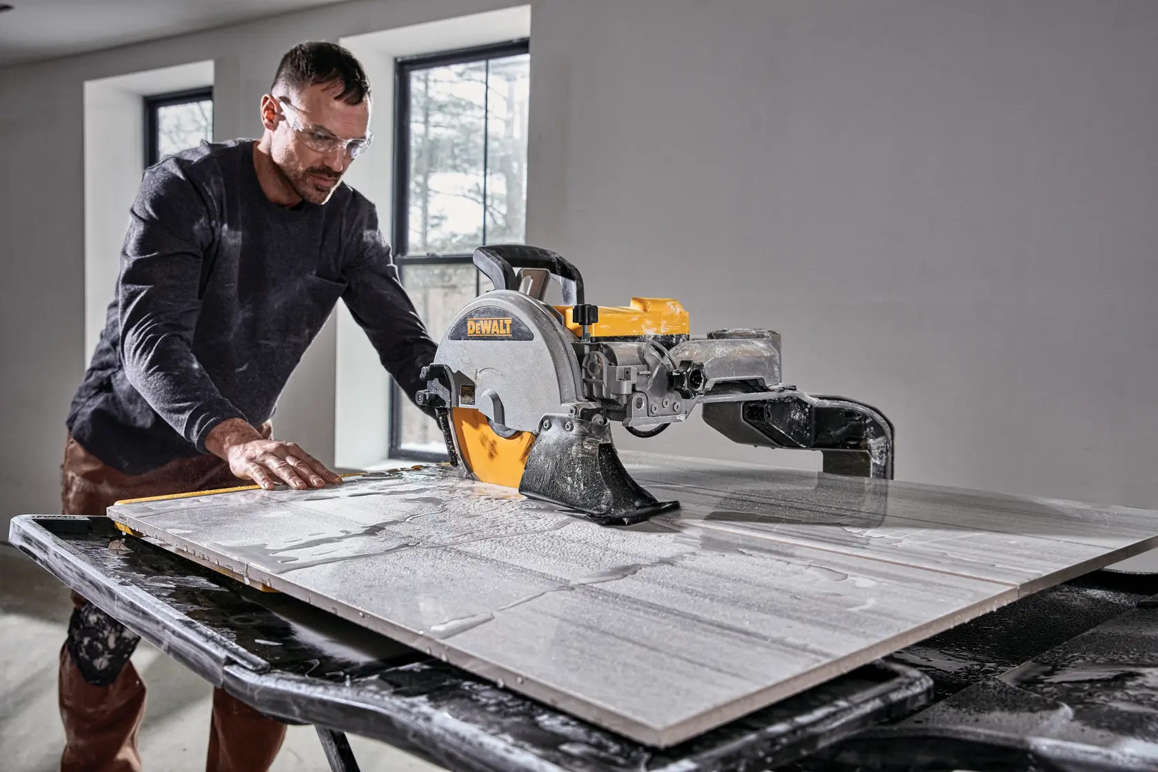 A person is operating a DEWALT D36000S wet tile saw, cutting a large tile on a worktable indoors. The individual's face is blurred.