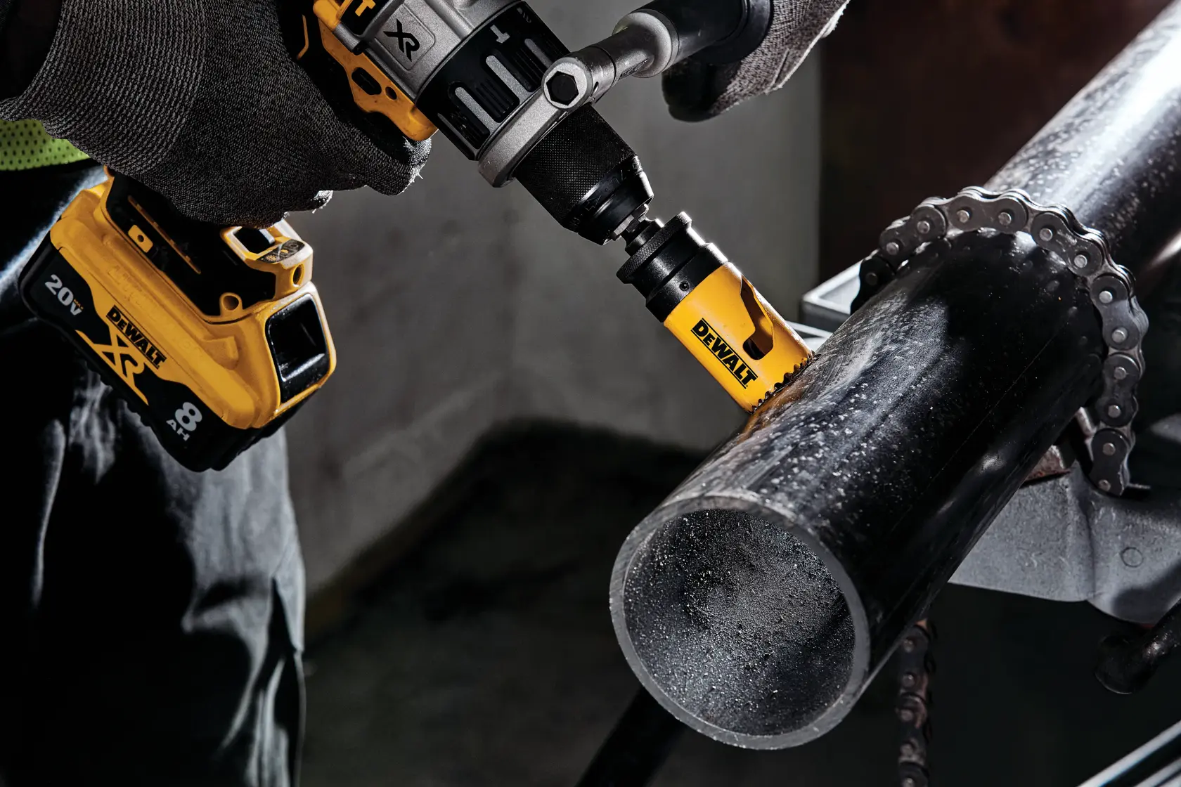 A close-up view of a person using a DEWALT cordless drill with a hole saw attachment to cut through a black metal pipe, with gloved hands holding the tool firmly.