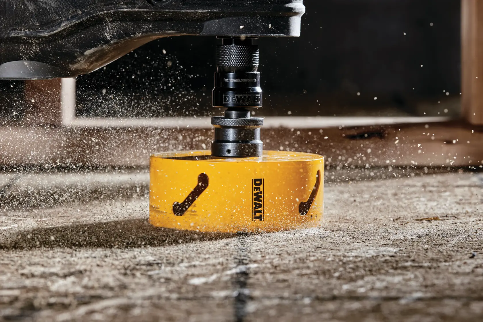 Close-up of a DEWALT yellow hole saw attachment cutting through a concrete surface, with dust and debris flying around.