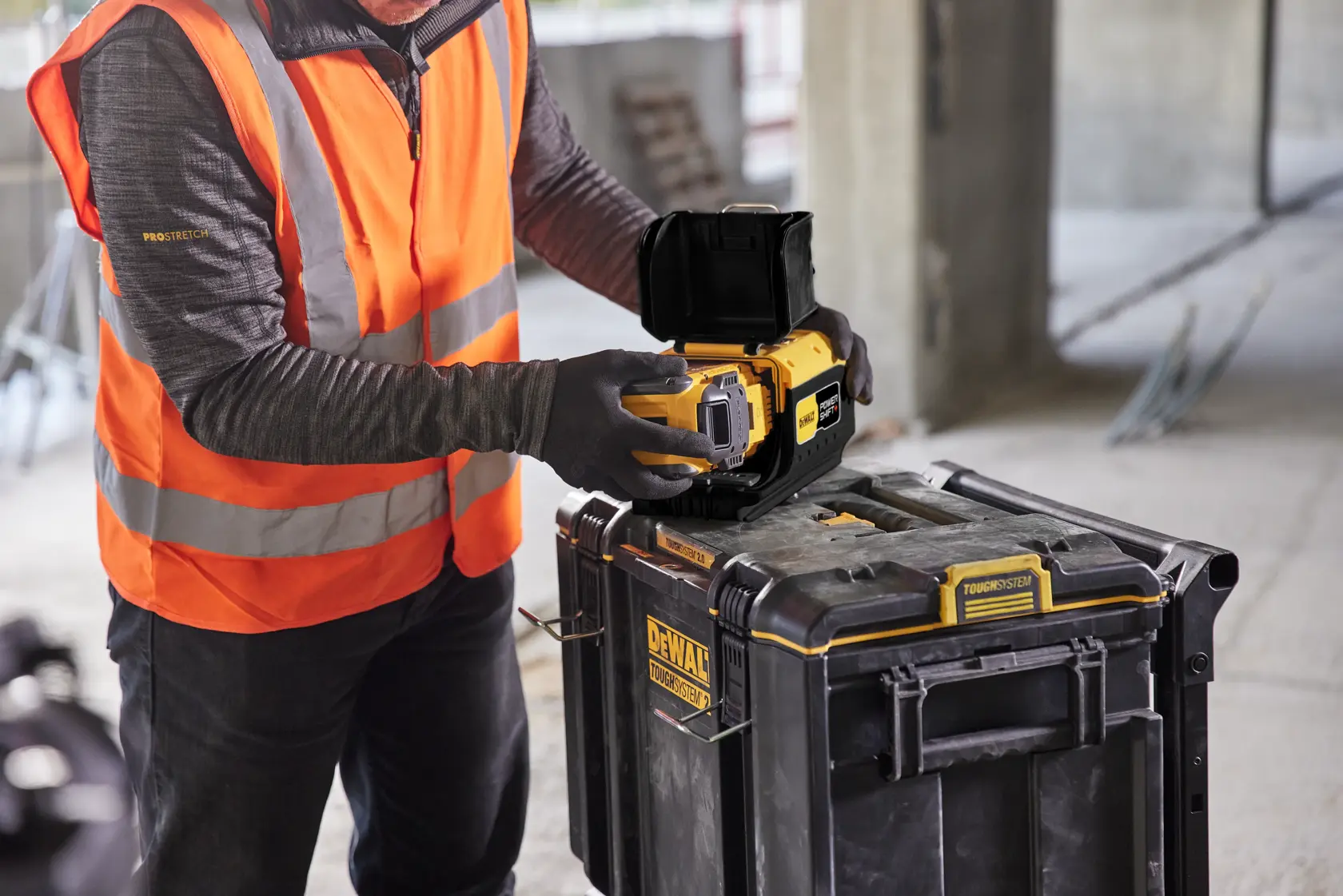 A person wearing an orange high-visibility vest and gloves is using a DEWALT device placed on a DEWALT ToughSystem storage box at a construction site.