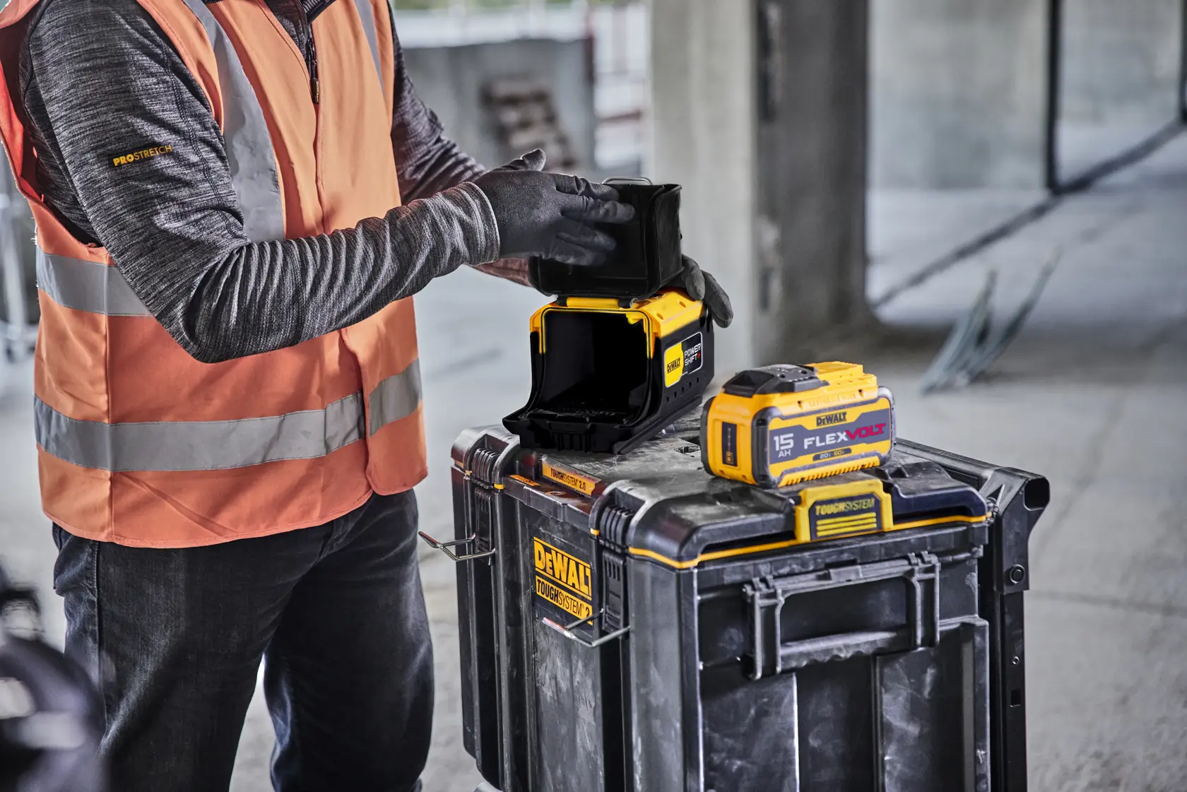 A person wearing an orange safety vest and gloves is handling a DEWALT photography product on top of a DEWALT ToughSystem storage case, with a DEWALT FLEXVOLT battery visible beside the product. The scene is set in an industrial or construction environment.