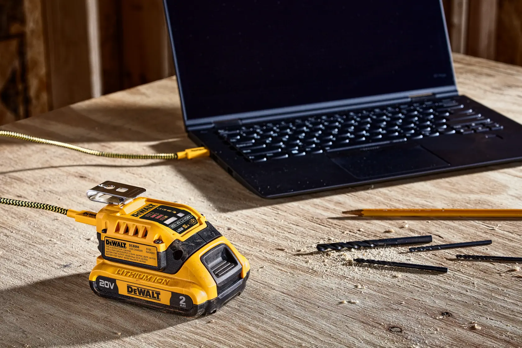 A DEWALT 20V lithium-ion battery with a USB adapter is charging a black laptop on a wooden workbench. There are also some pencils and drill bits scattered on the table.