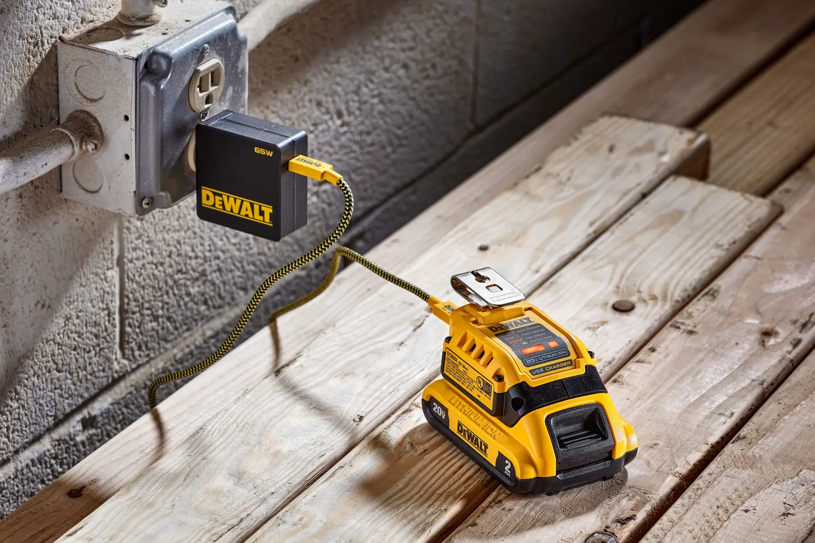 A DEWALT portable battery charger kit is plugged into a wall electrical outlet with a yellow and black cable, resting on a wooden surface. The kit includes the DEWALT battery charger and adapter, suitable for power tools.