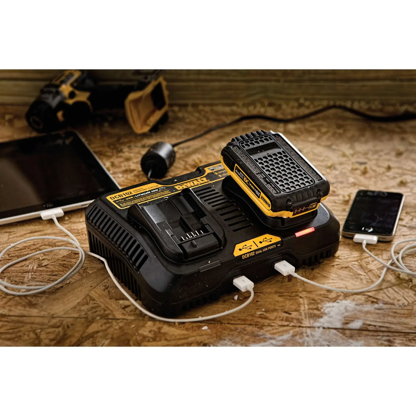 A DEWALT DCB102 dual battery charger with a battery mounted on top, charging two electronic devices using USB ports, on a wooden work surface. A tablet and a smartphone are connected to the charger, and a DEWALT power tool is visible in the background.