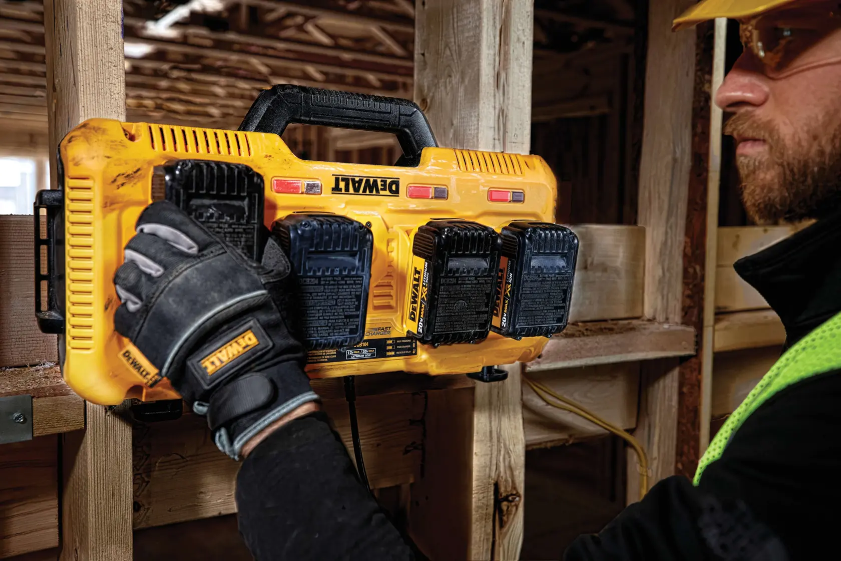 A yellow DEWALT four-port battery charger on a wooden floor, charging four DEWALT batteries. The charger supports 12V Max, 20V Max, and FLEXVOLT 20V/60V Max batteries. Nearby are construction materials and a white cabinet door.