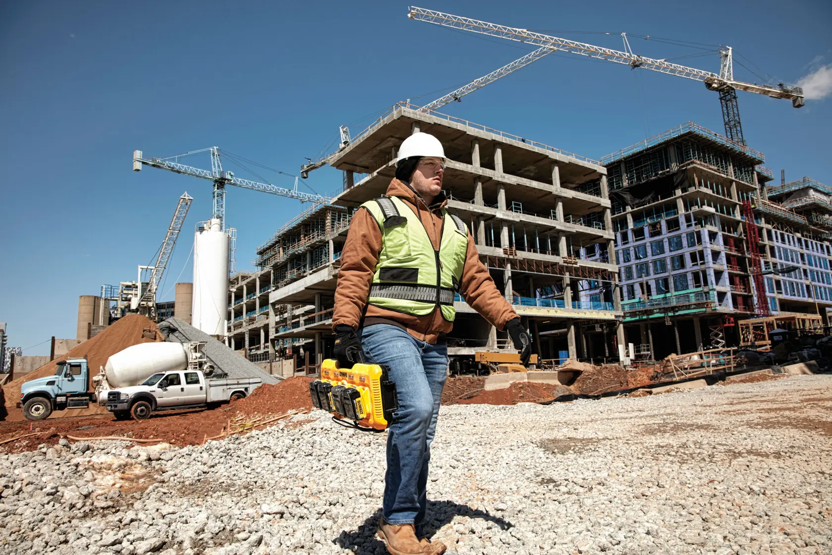 A construction worker wearing a safety vest, jacket, helmet, and gloves is walking at a construction site carrying a DEWALT DCB104 battery charger with four batteries attached. The background shows multi-story building structures under construction with cranes and construction vehicles. Text on the image highlights fast charging times for DCB204 and DCB606 batteries.