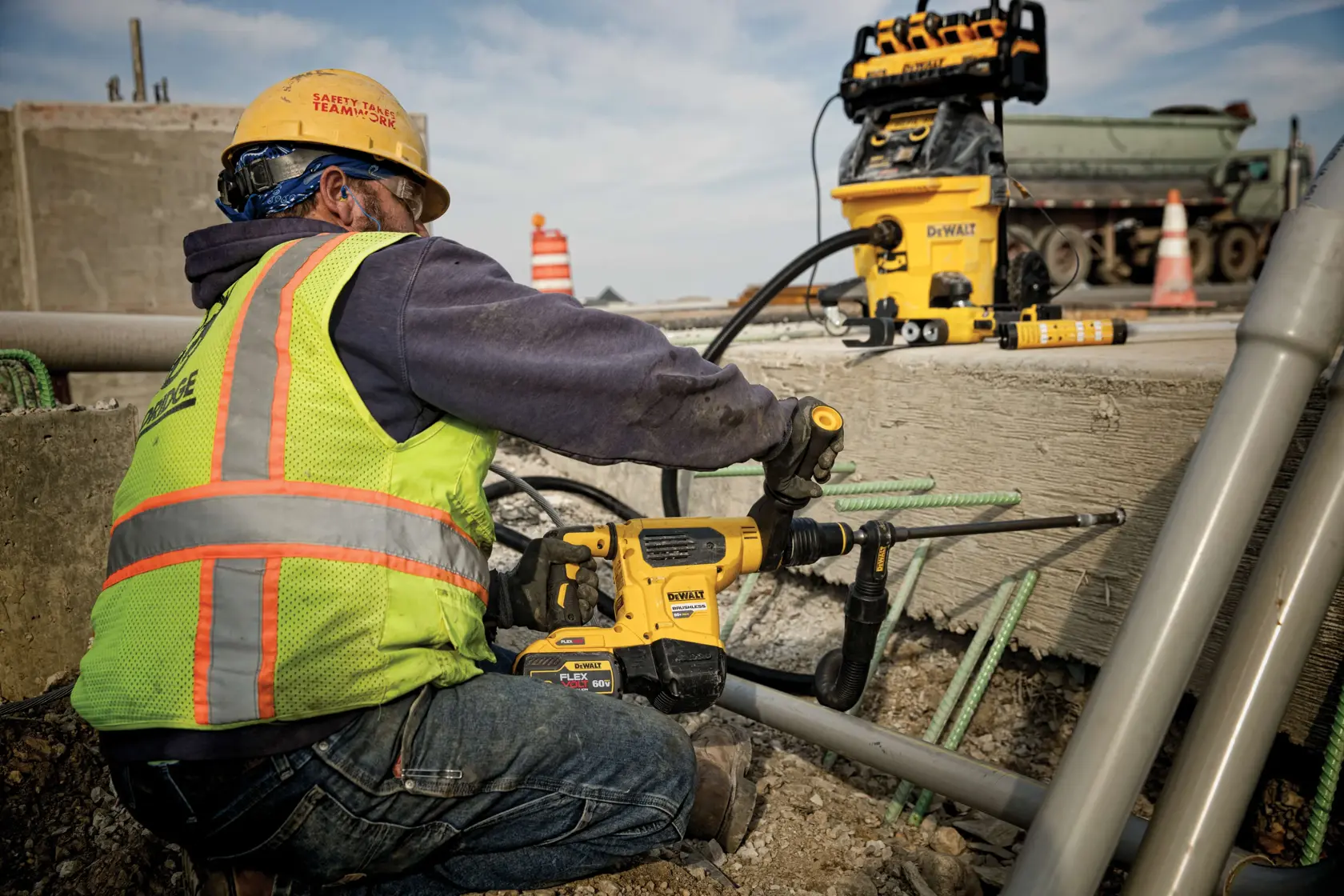 A worker wearing a safety vest and helmet operates a DEWALT power tool to drill into concrete at a construction site. Other DEWALT equipment and construction materials are visible in the background.