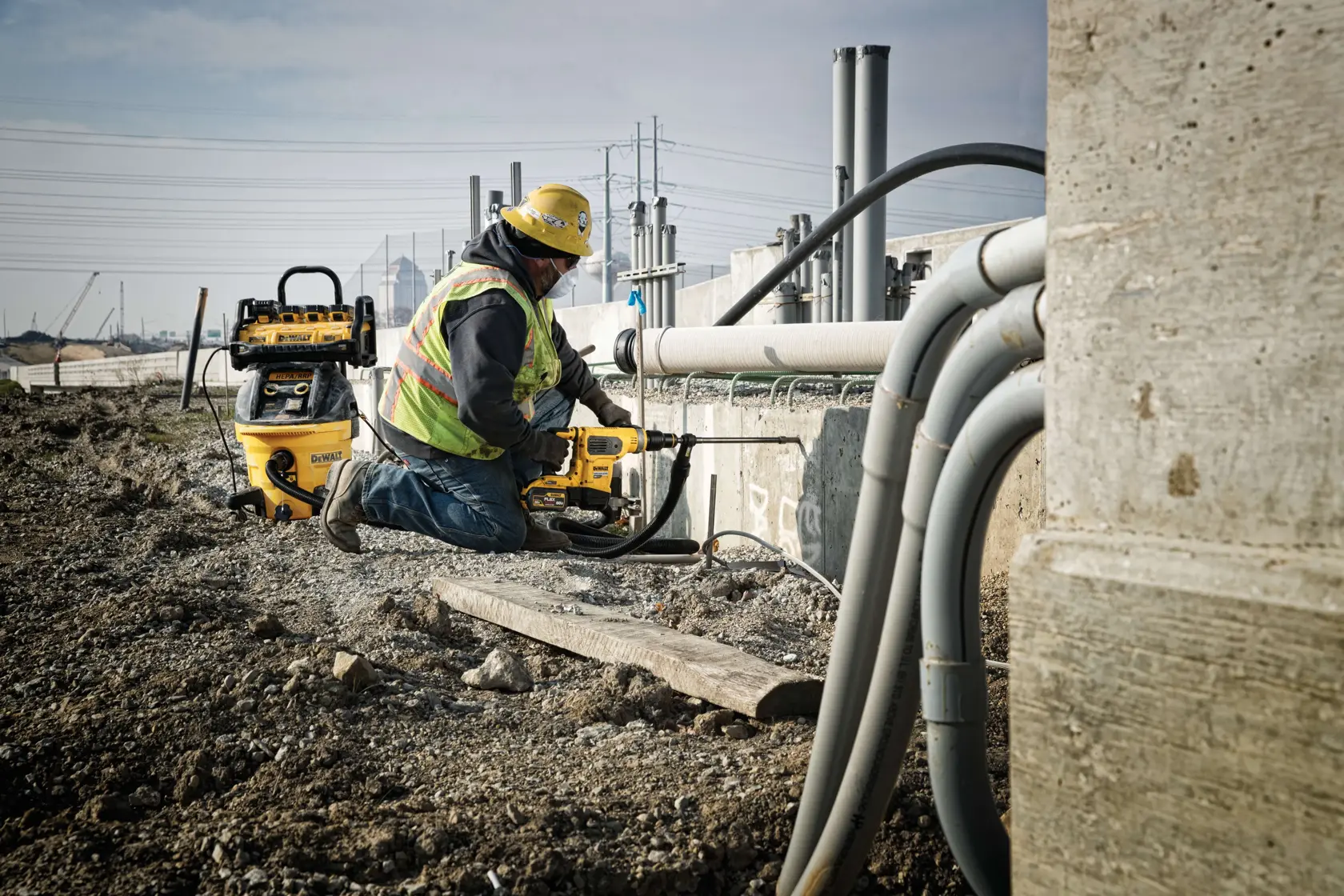 A construction worker wearing a hard hat and safety vest is kneeling on the ground, using a DEWALT power tool near a building site with exposed pipes and concrete. Several DEWALT tools and equipment are visible in the background.