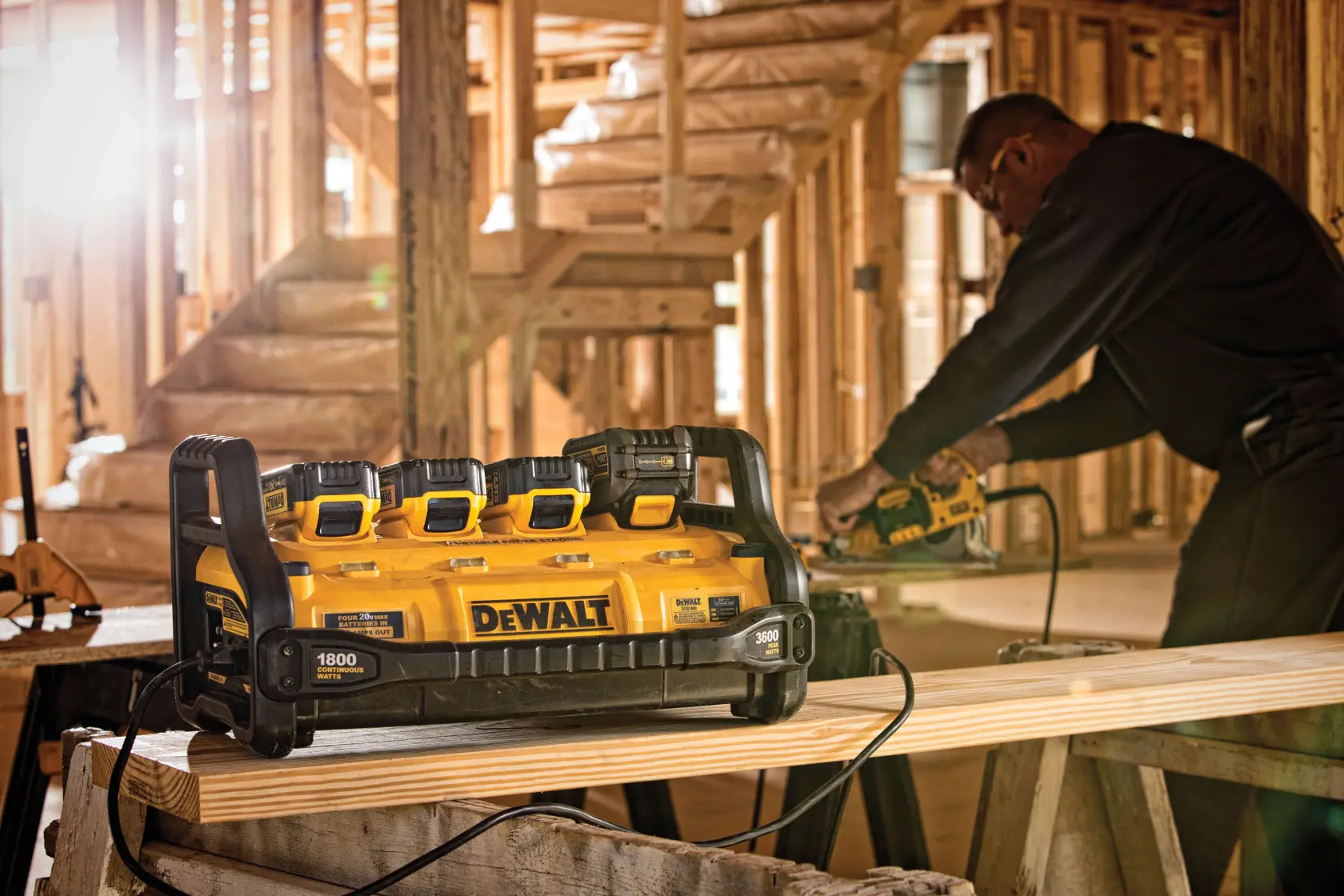 A DEWALT power station with four battery slots is placed on a wooden plank in a construction site. In the background, a person is using a DEWALT power tool to work on the wood.