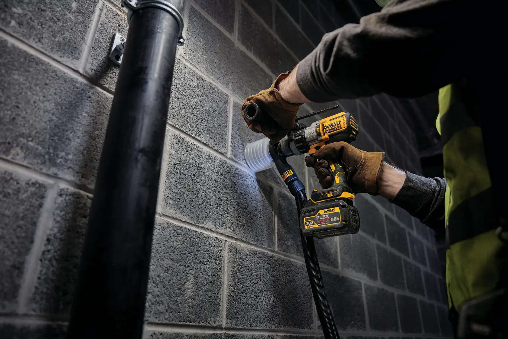 A person wearing work gloves and a safety vest is using a DEWALT cordless drill with a DCB606 FLEXVOLT battery to drill into a concrete block wall, with a dust extraction attachment connected.