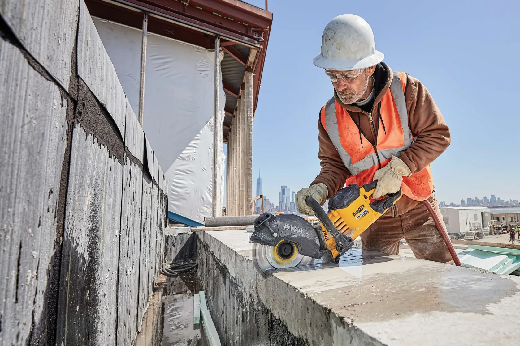Worker uses a DEWALT power tool to cut concrete at an outdoor construction site, wearing safety gear and helmet.