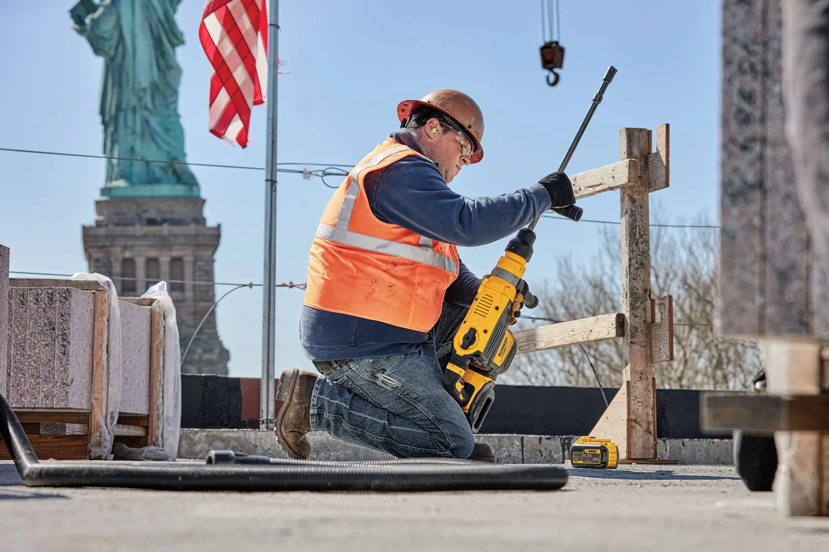 A construction worker wearing a hard hat and orange safety vest is using a yellow DEWALT power tool to drill into a concrete surface at a job site. Granite blocks and construction materials are visible nearby, with the Statue of Liberty and an American flag in the background.