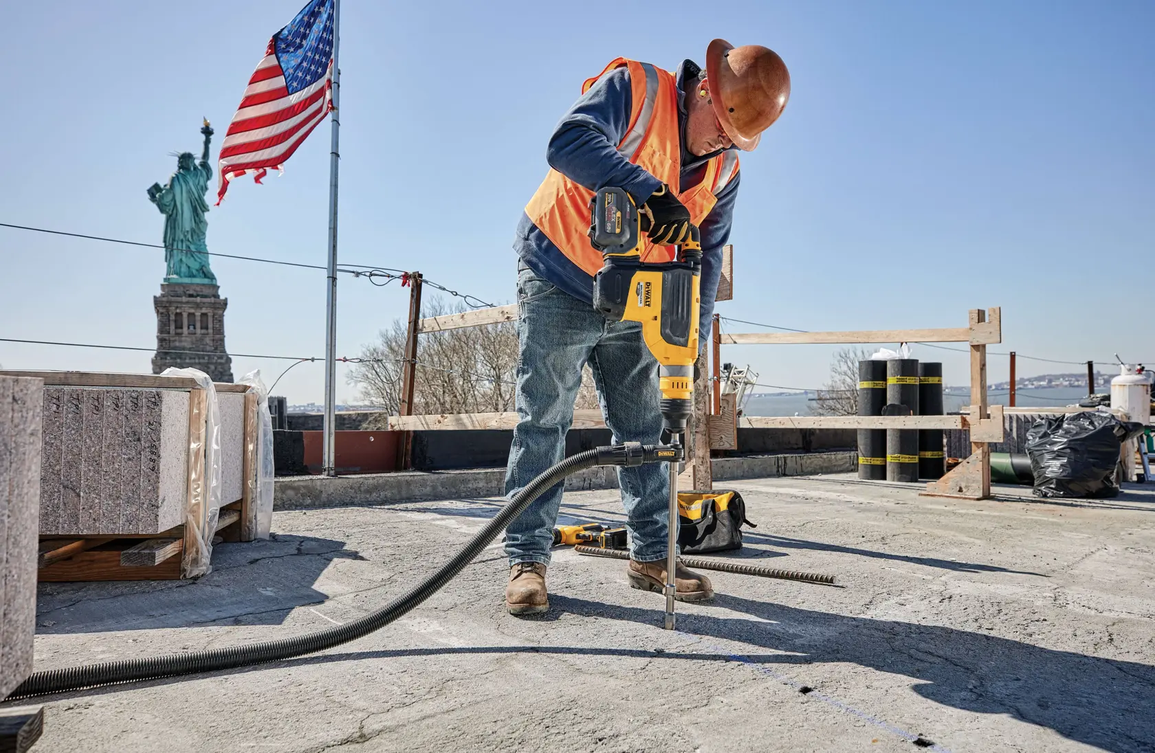 A construction worker wearing a safety vest, gloves, and a helmet is using a DEWALT power tool to cut concrete on a construction site. The worker’s face is blurred, and the DEWALT tool is clearly visible. The background shows ongoing construction and a clear sky.