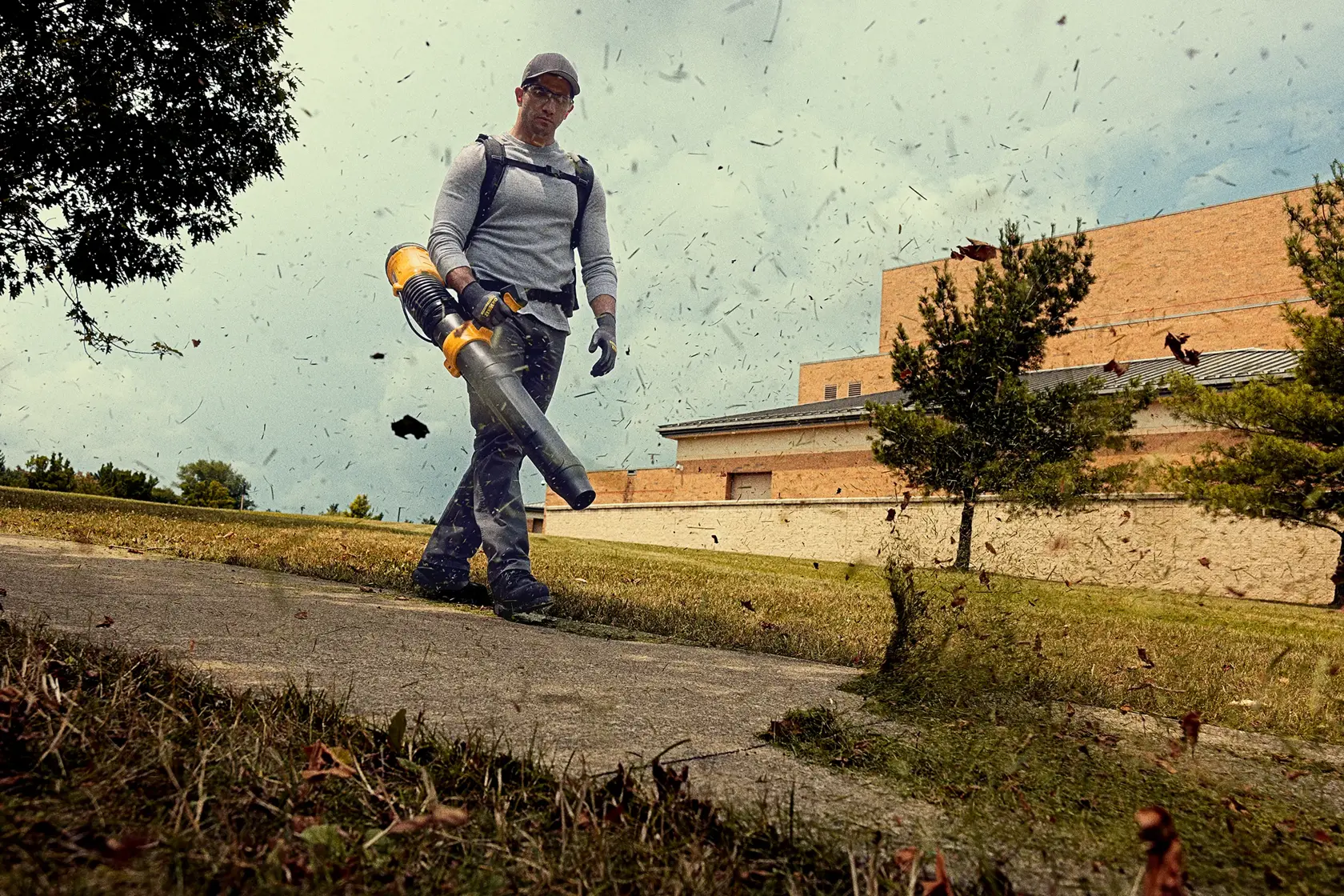 A person wearing gloves operates a DEWALT cordless leaf blower, walking along a sidewalk in an outdoor area with grass, trees, and a building in the background. Leaves and debris are being blown through the air.